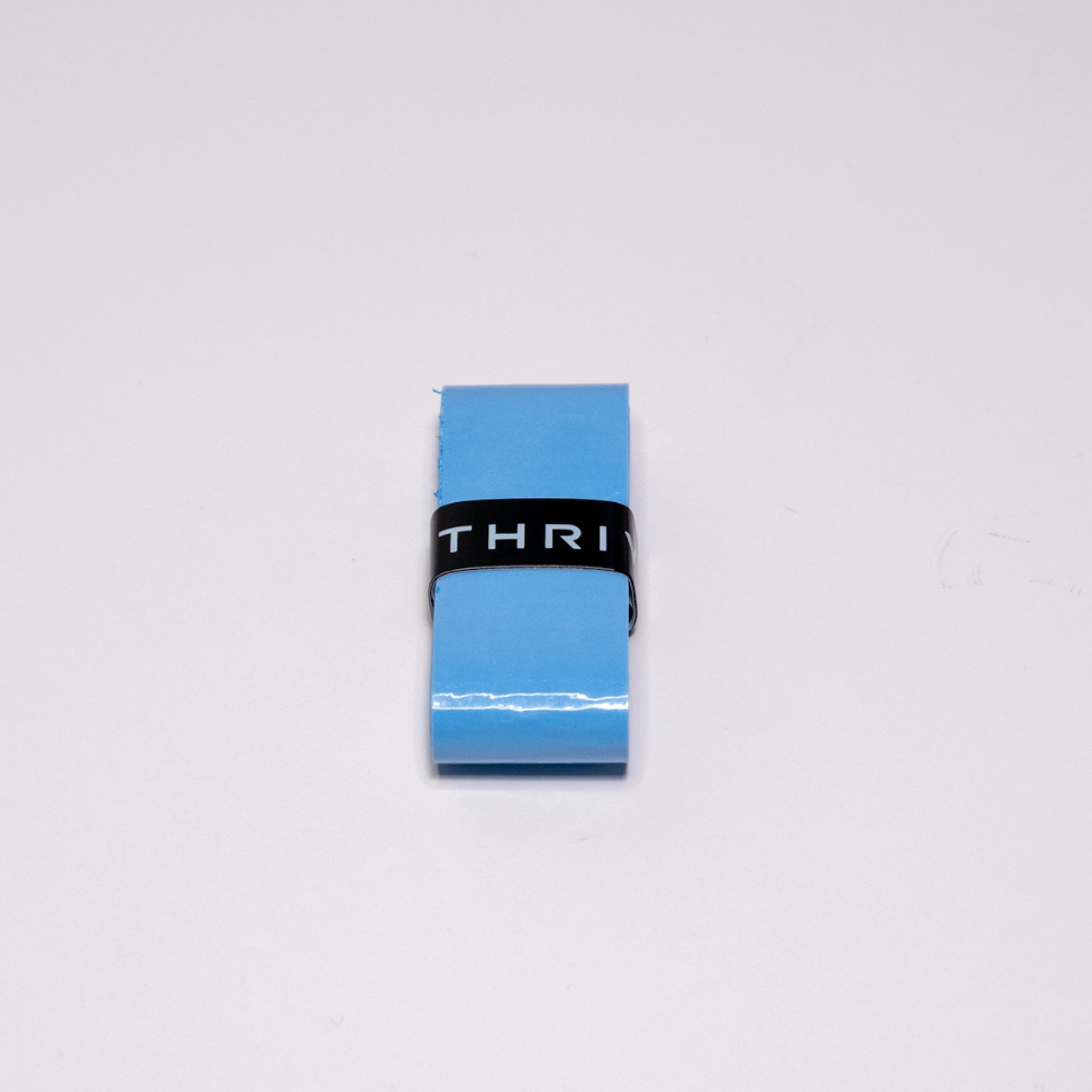A rolled-up blue Thrive Pickleball Overgrips (Super Tacky) sits centered on a white surface, secured by a black strap featuring partially visible "THRI" text.