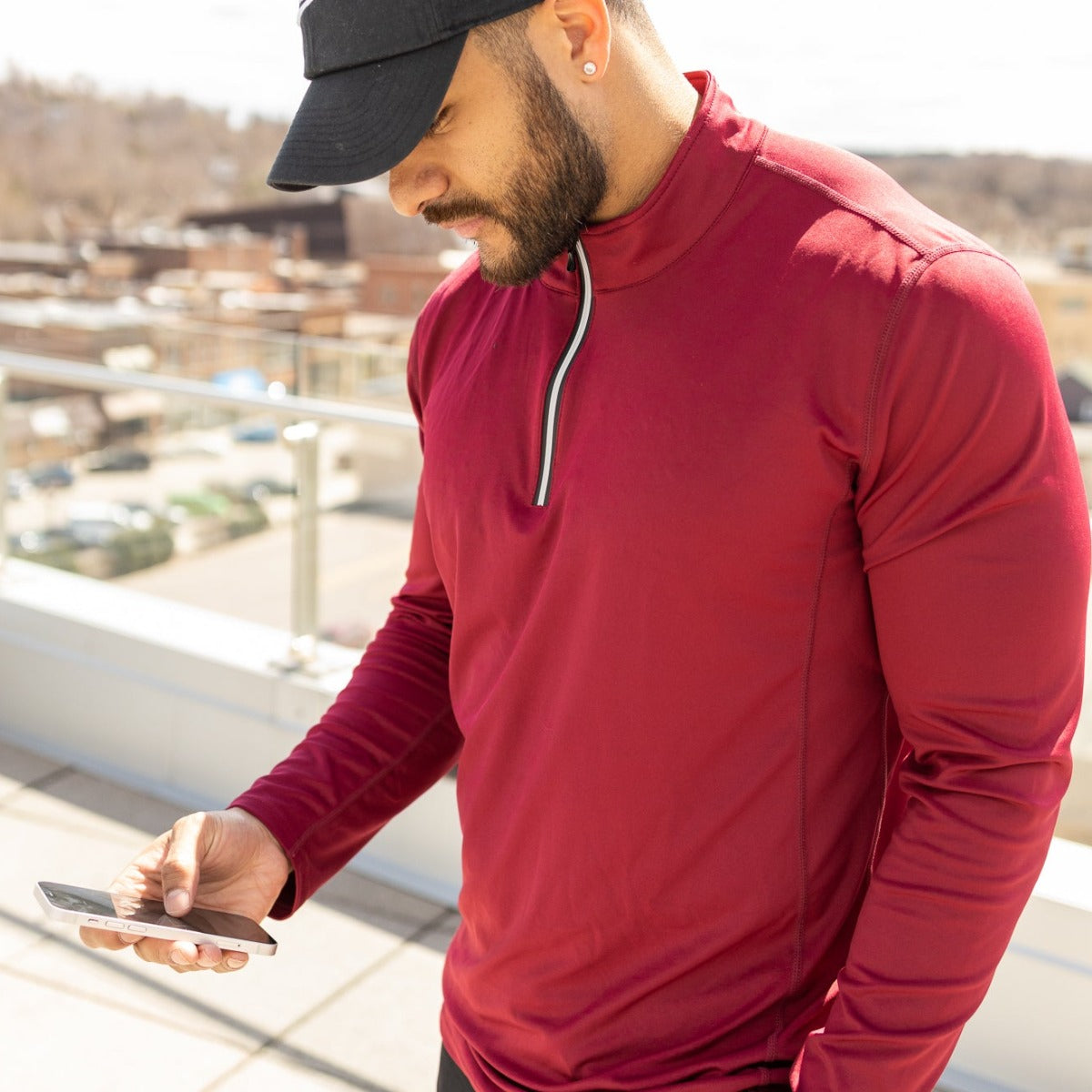 A man outdoors in DYM Athletics FLEX Men's 1/4 Zip (SALE), paired with a black cap and stud earring, looks down at his smartphone. Buildings, trees, and glass-paneled railings form the background.