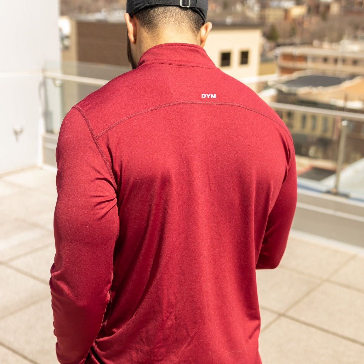 A man in a black cap and a DYM Athletics FLEX Men's 1/4 Zip red long-sleeve stands on a sunlit rooftop with glass railings and city buildings in the background.