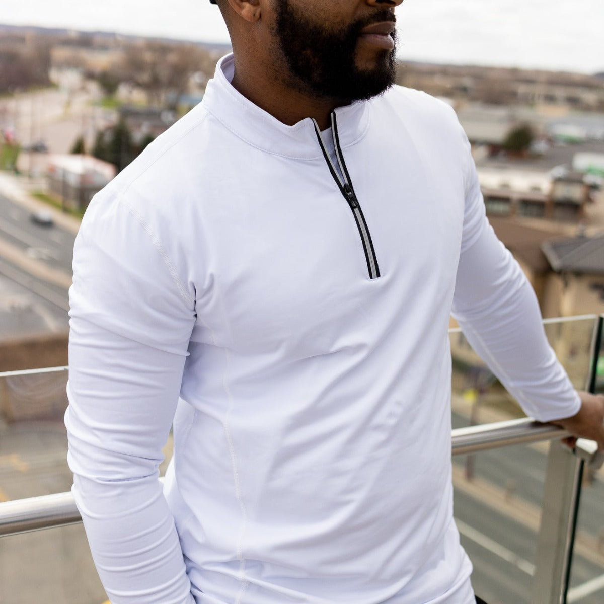 A man stands on a balcony above a city street, wearing the DYM Athletics FLEX Men's 1/4 Zip pullover in white—perfect for men's activewear. His face is partially out of frame, with blurred buildings and roads behind him.