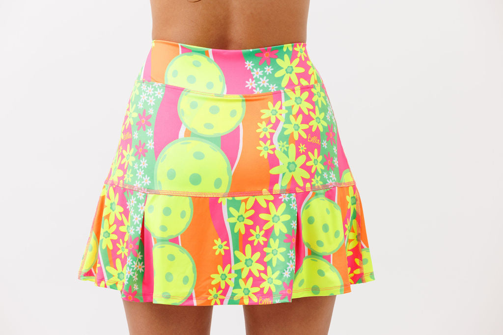 A person models the Pickleball Bella PB Drop Pleat Skort Daisy featuring a wide waistband and pleated lower section with green pickleballs and yellow daisies on a pink, orange, and green background against a white backdrop.