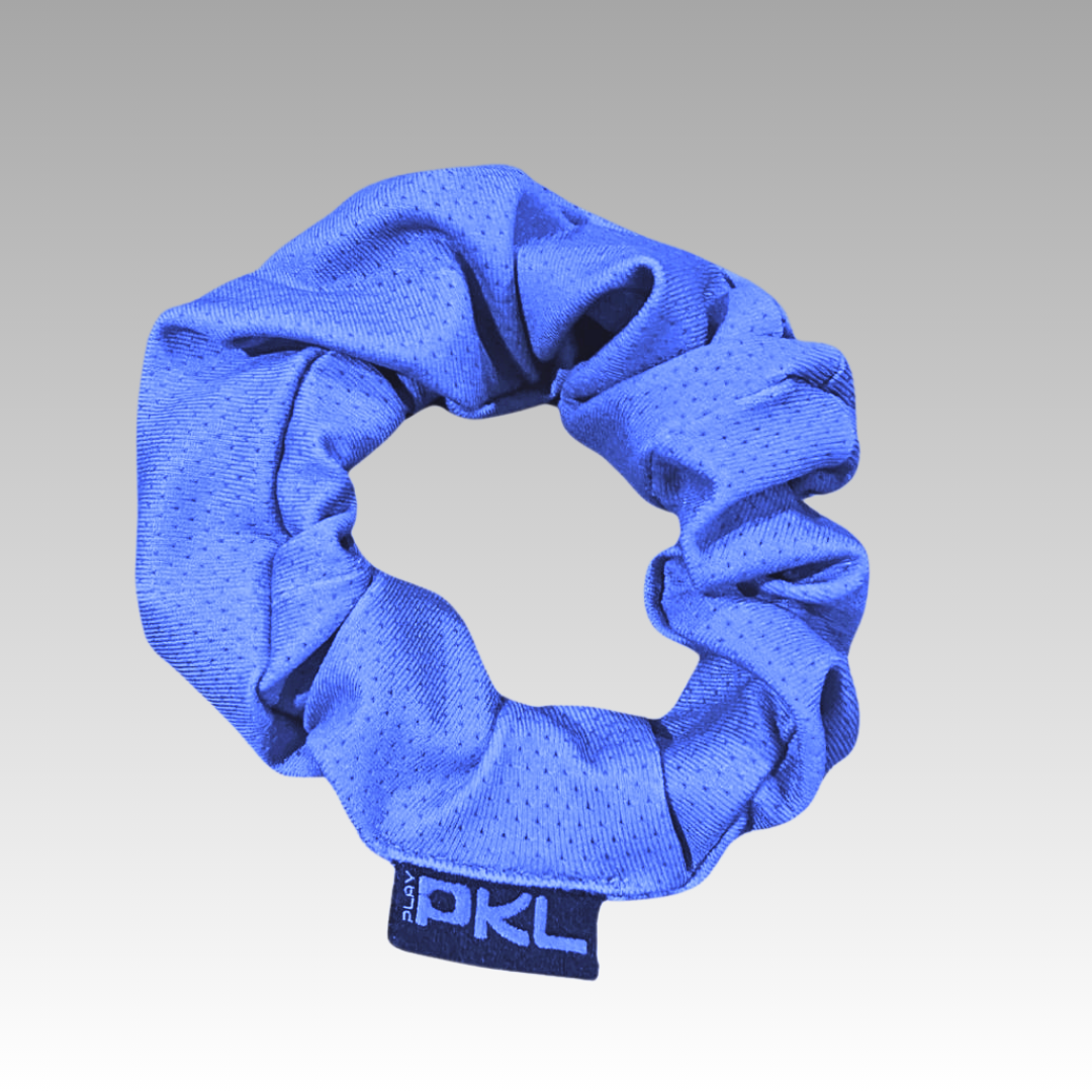 The Shanti Pickleball Hair Scrunchie by Playly Store is a blue fabric accessory with a subtle dotted pattern and a small black tag with white "PKL" text, displayed neatly in a circular shape against a gray gradient background.