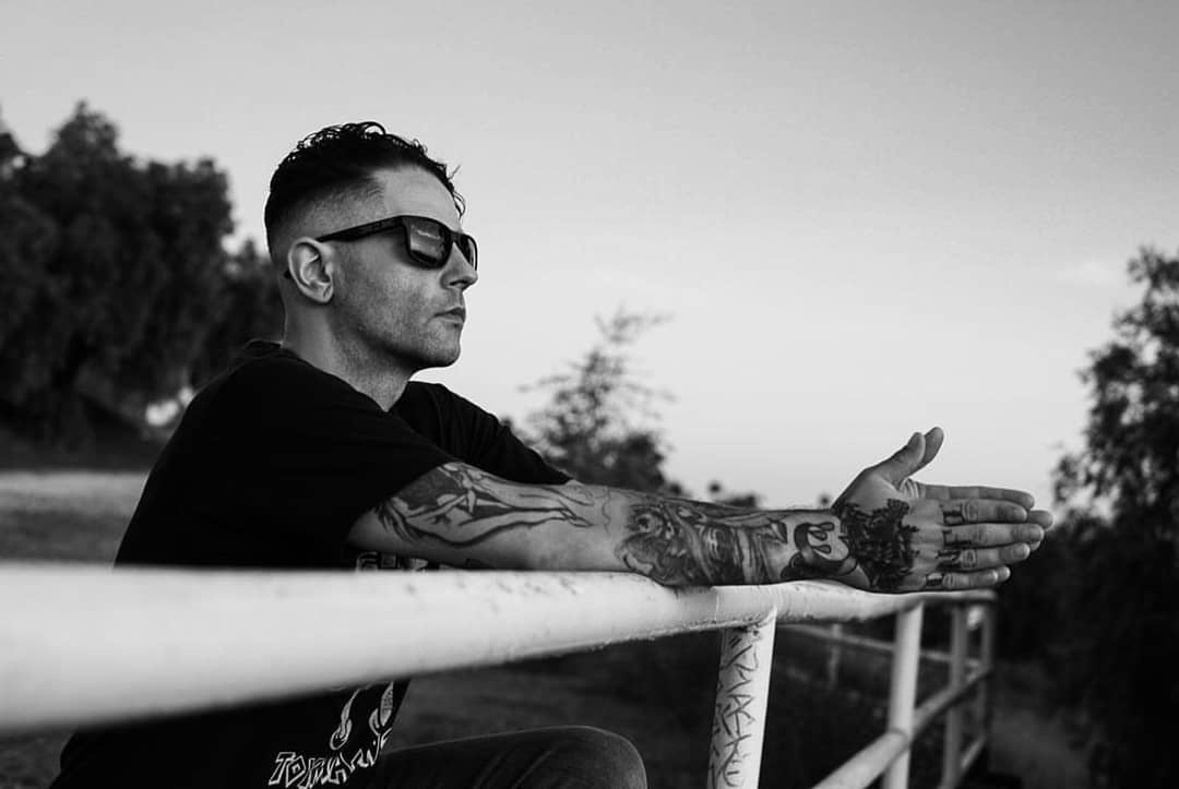 Wearing Epoch Eyewear’s Delta sunglasses, a tattooed man relaxes outside, arms on a metal railing and eyes set on the horizon—a black and white scene framed by trees highlights his reflective mood under the clear sky.