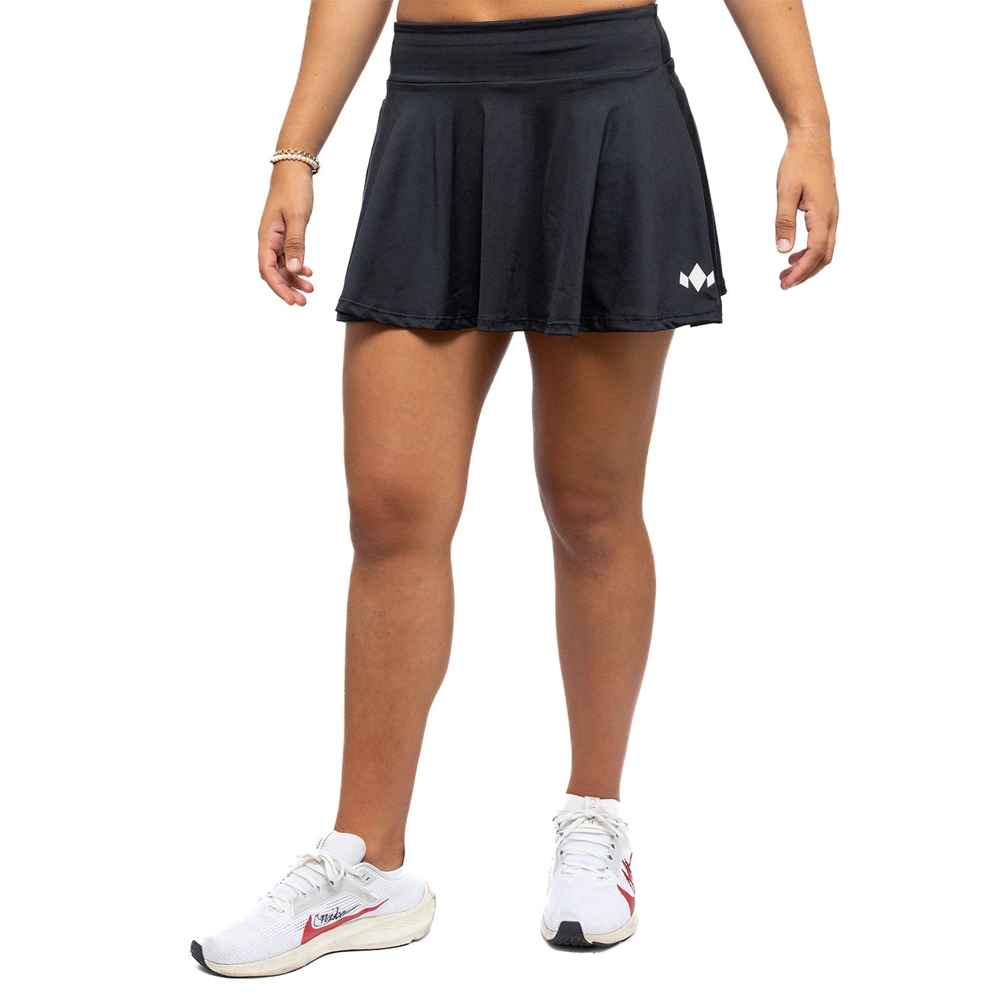 A person models the Diadem Sports Women's Essential Tennis and Club Performance Skirt—a short black skirt with a small white logo—paired with white socks and white running shoes with red accents, shown against a plain white background.