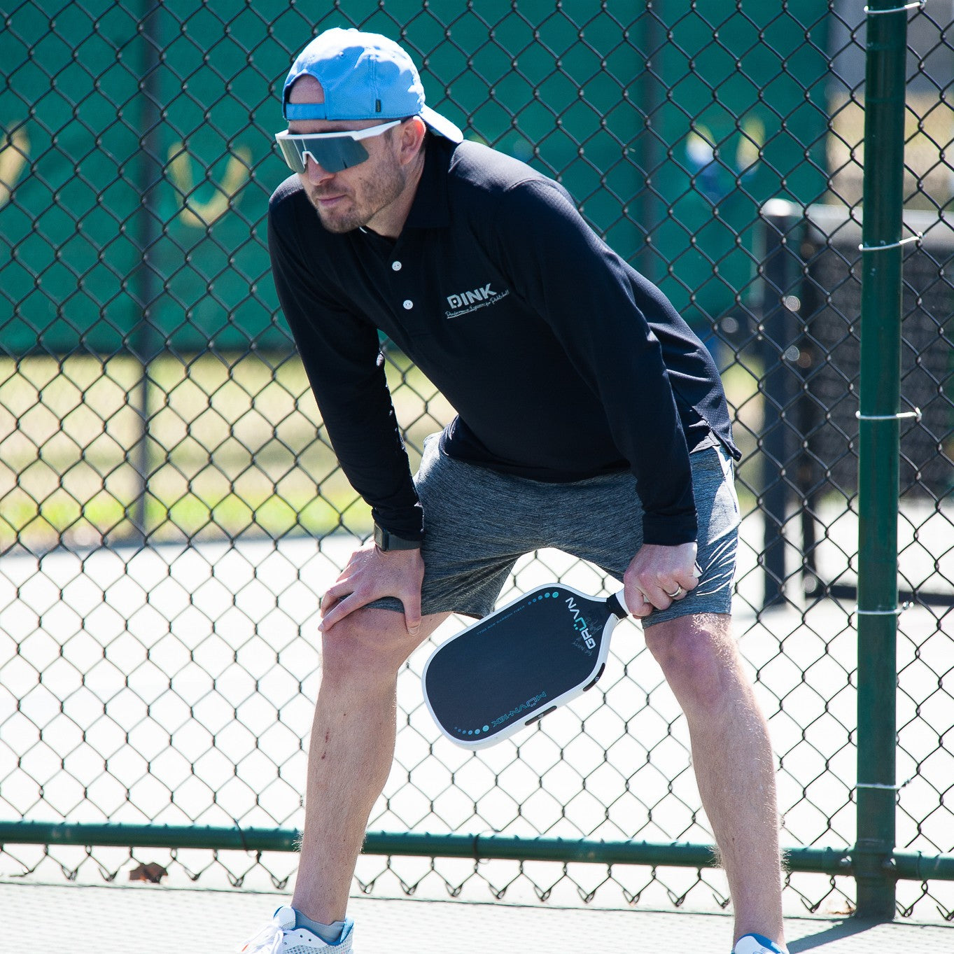 A man in a blue cap, black long-sleeve shirt, and gray shorts grips a pickleball paddle on an outdoor court. He wears Dink Eyewear Captiva Large White Pearl Pickleball Sunglasses, with sunlight and greenery behind him by a chain-link fence.