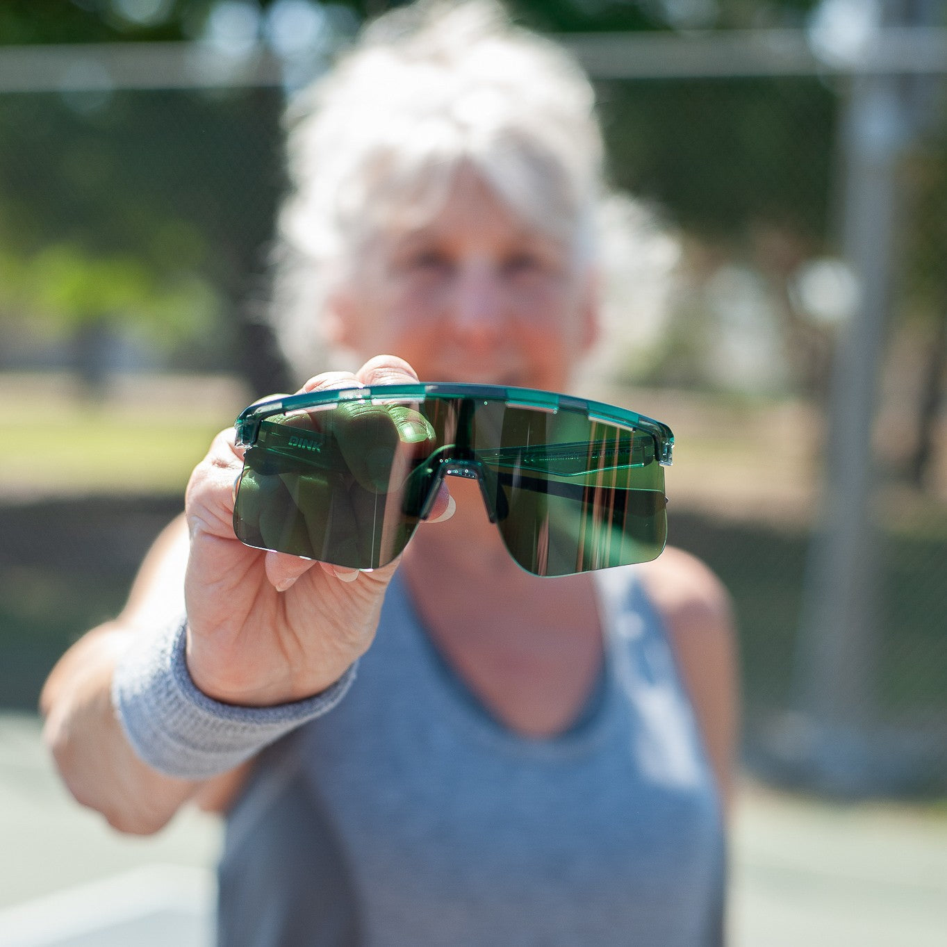 An older woman in a gray tank top stands outdoors on a sunny day, holding Captiva Emerald Green Sunglasses from Dink Eyewear with ColorBoost lens technology toward the camera. Her face is softly blurred; a tennis court and trees are visible behind her.