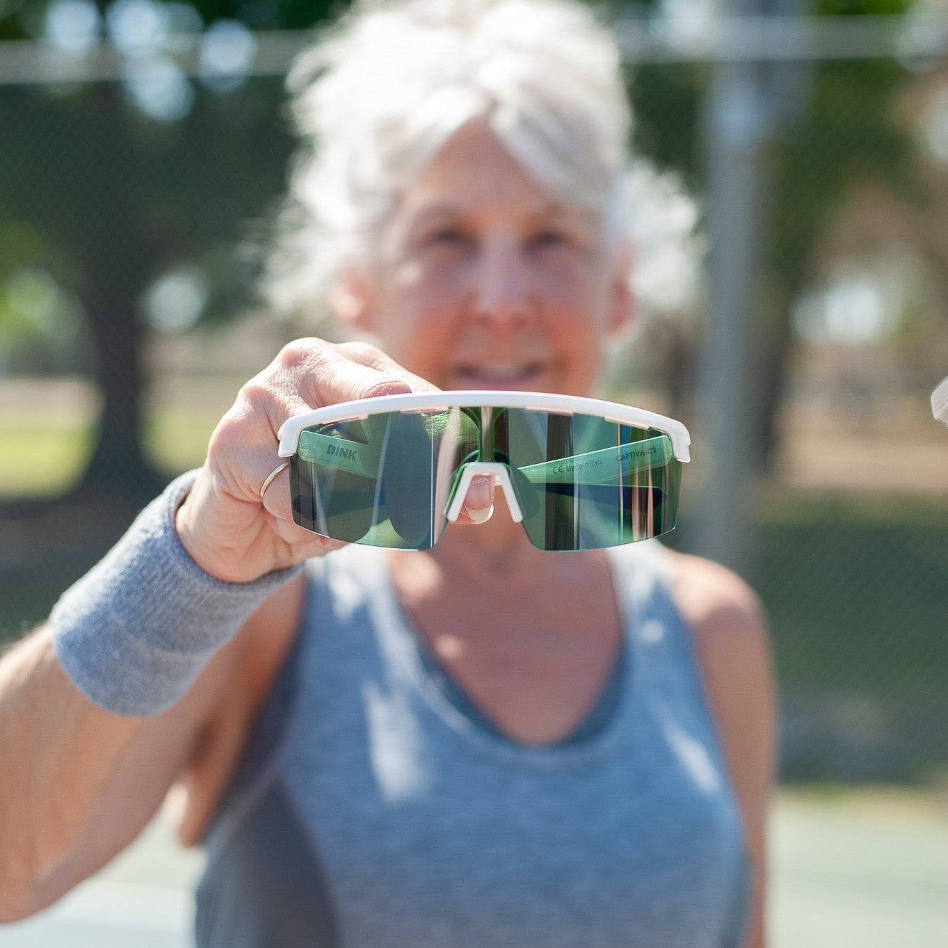 An older woman with gray hair in a ponytail, wearing a gray tank top and wristband, holds Dink Eyewear Captiva Pickleball Sunglasses with ColorBoost lens technology and UV protection toward the camera on a sunny outdoor court.