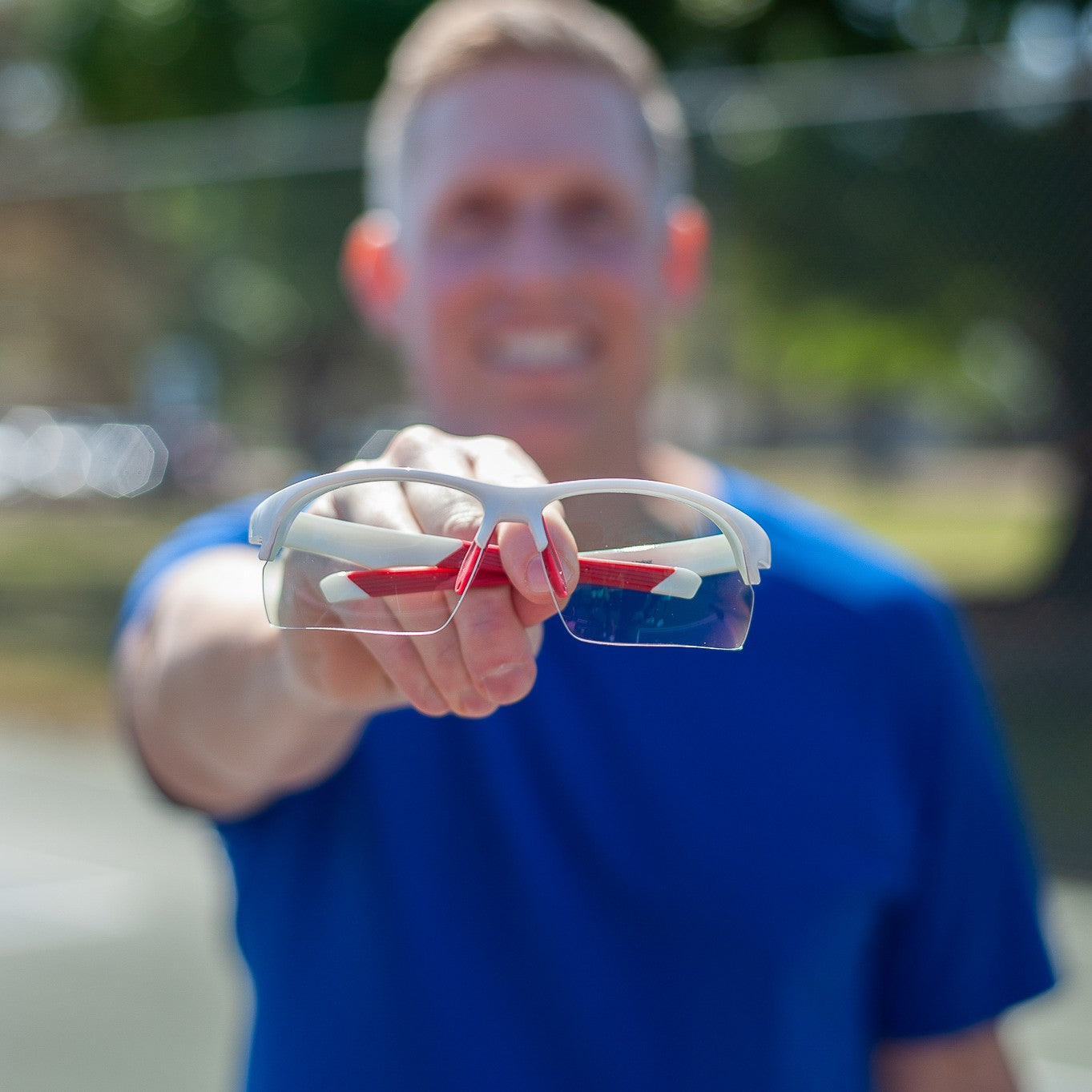 A person in a blue shirt stands outdoors on a sunny day, holding Dink Eyewear’s Daytona Pickleball Sunglasses - White/Red in a sporty wrap design toward the camera. The sunglasses are in sharp focus; the face and trees behind are blurred.