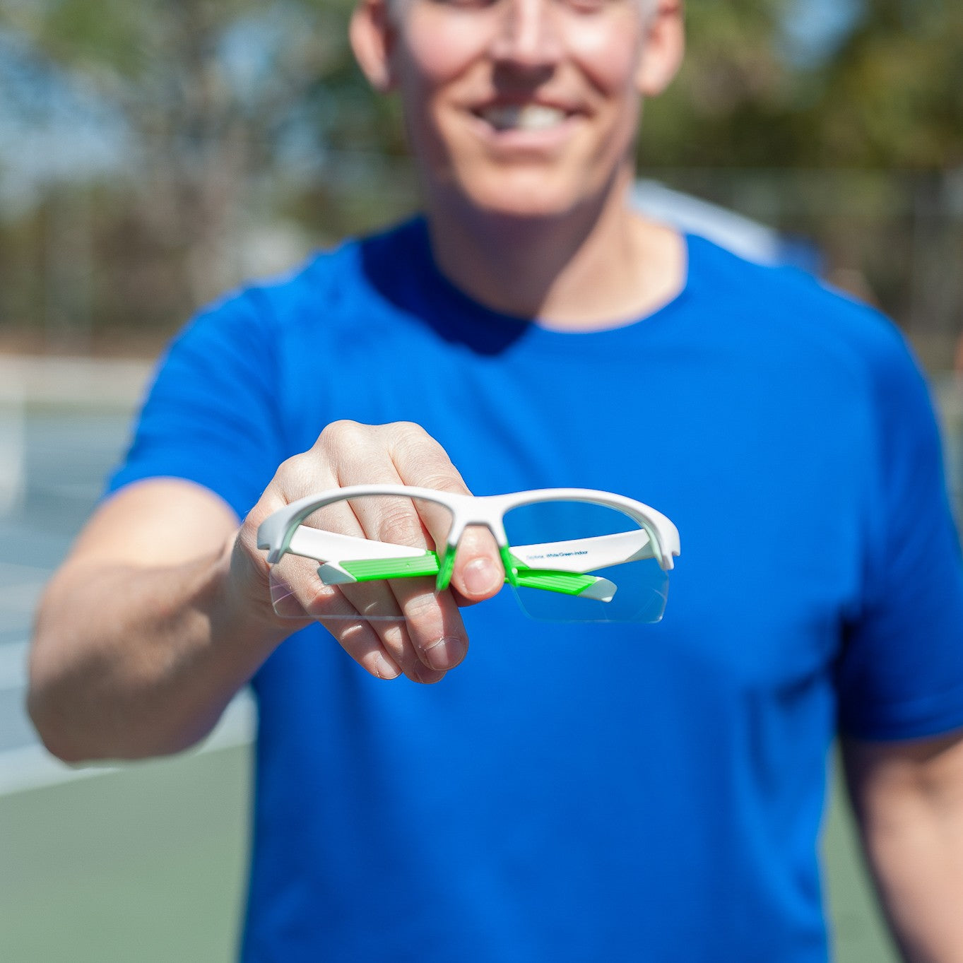 A smiling person in a bright blue shirt holds Dink Eyewear's Daytona Sunglasses (White/Green) toward the camera outdoors on a sunny day, with a blurred tennis court and trees behind them. The focus is on the sunglasses, not the person's face.