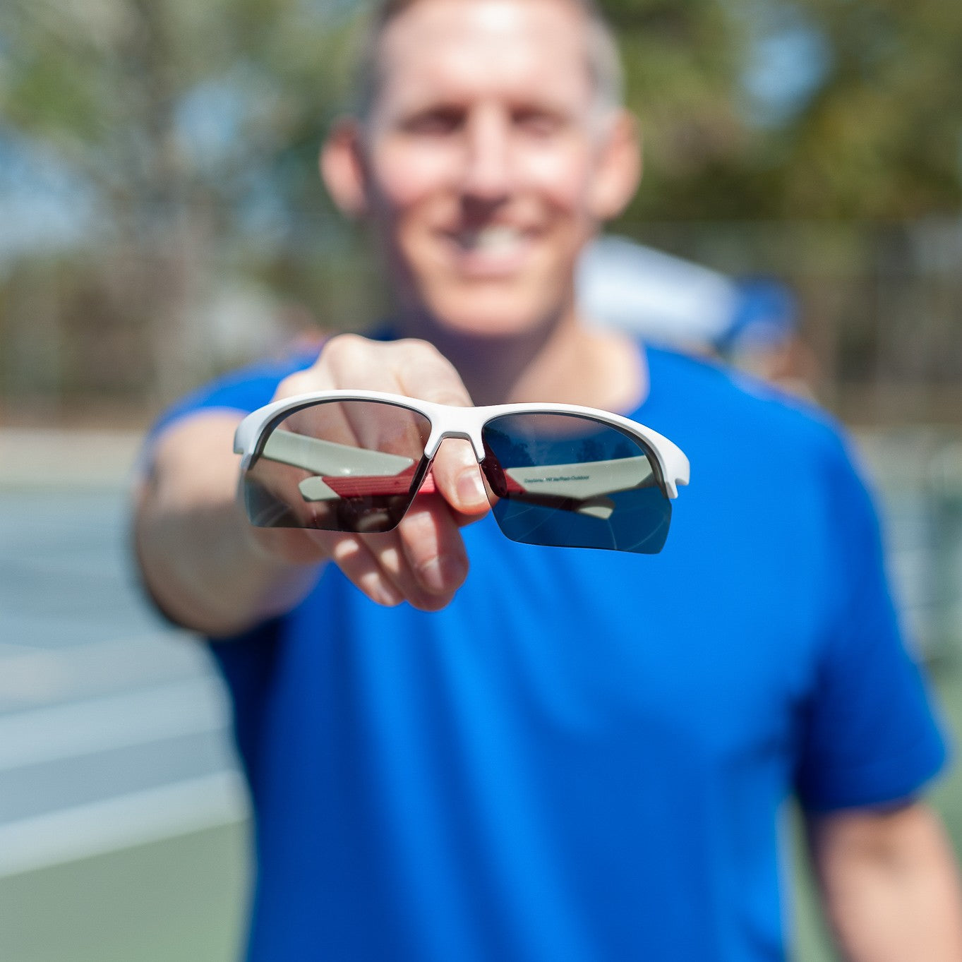 A man in a blue shirt stands on a tennis court, extending Dink Eyewear's Daytona Pickleball Sunglasses (White/Red) toward the camera. The sharp focus highlights the stylish frames, while his face and the background are blurred.