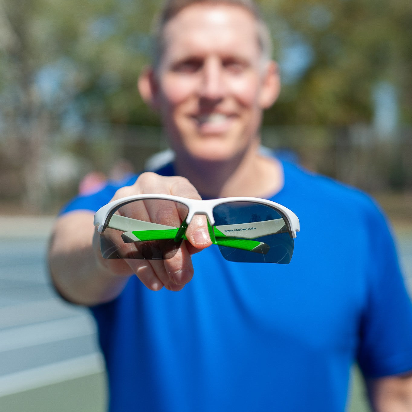 A person in a bright blue shirt holds Dink Eyewear Daytona Pickleball Sunglasses in White/Green toward the camera. The sporty wrap-around lenses reflect tennis court lines, with trees and a blurred court in the background.