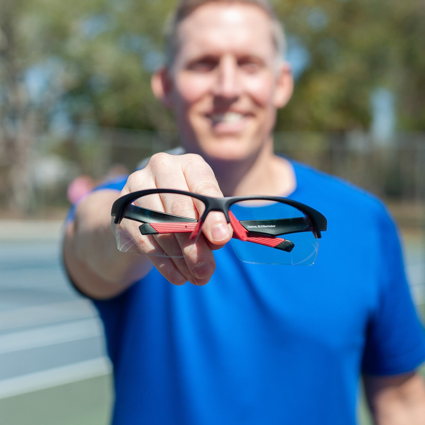 A smiling person in a blue shirt stands on a tennis court, holding up Dink Eyewear's Daytona Pickleball Sunglasses in Black/Red toward the camera, with the background blurred.