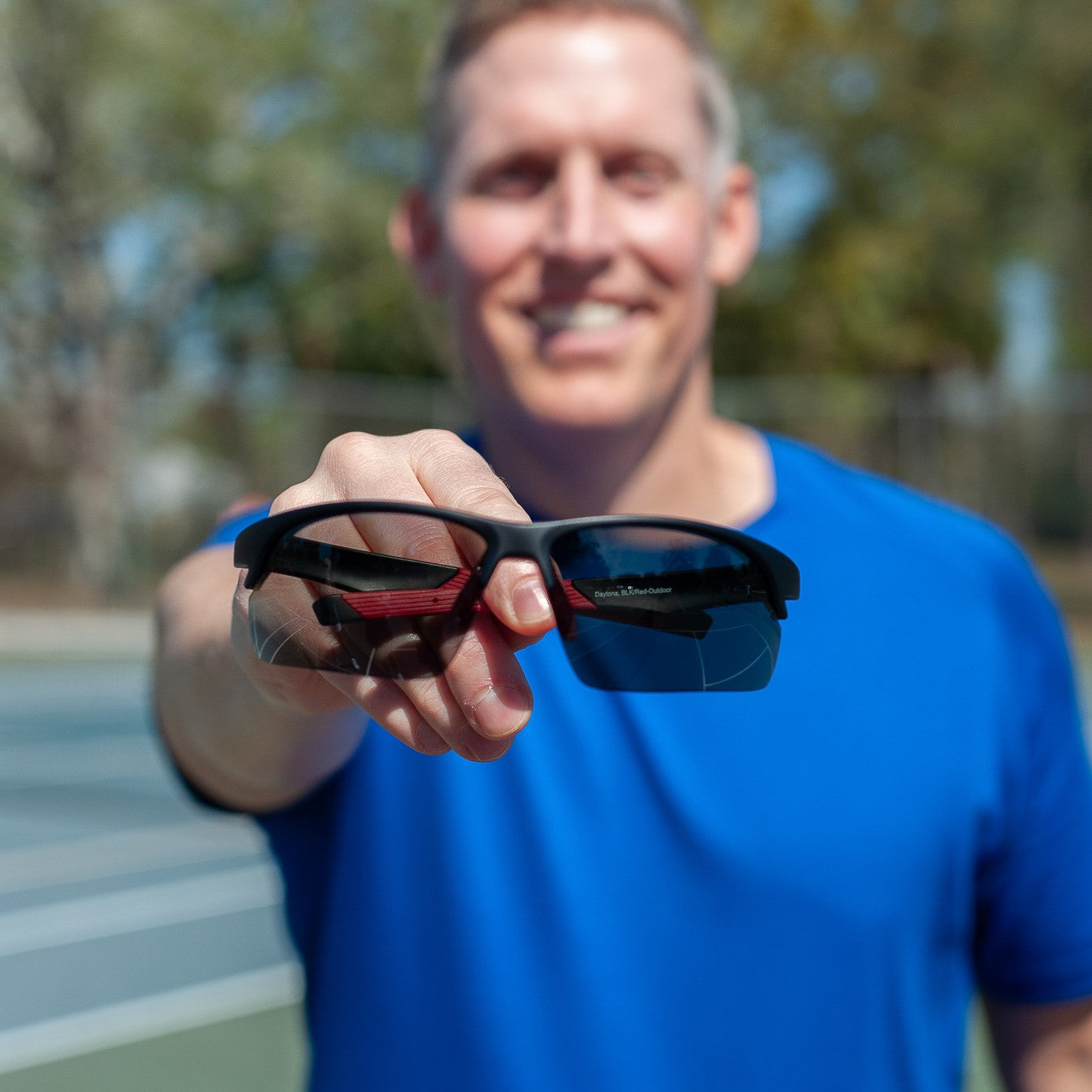 A smiling man in a blue shirt stands on an outdoor tennis court, holding Dink Eyewear’s Daytona Sunglasses - Black/Red with a sporty wrap design toward the camera, as trees and a fence appear blurred in the background.
