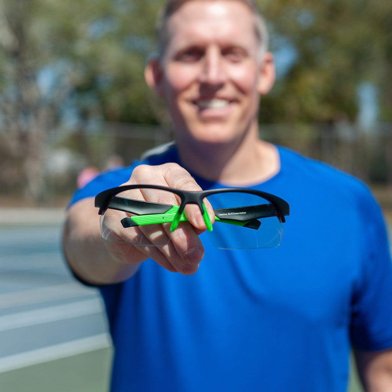 A smiling man in a blue shirt stands on a tennis court, holding out Dink Eyewear’s Daytona Sunglasses - Black Green toward the camera. The lightweight frame is in focus, while trees and a fence appear blurred in the background.