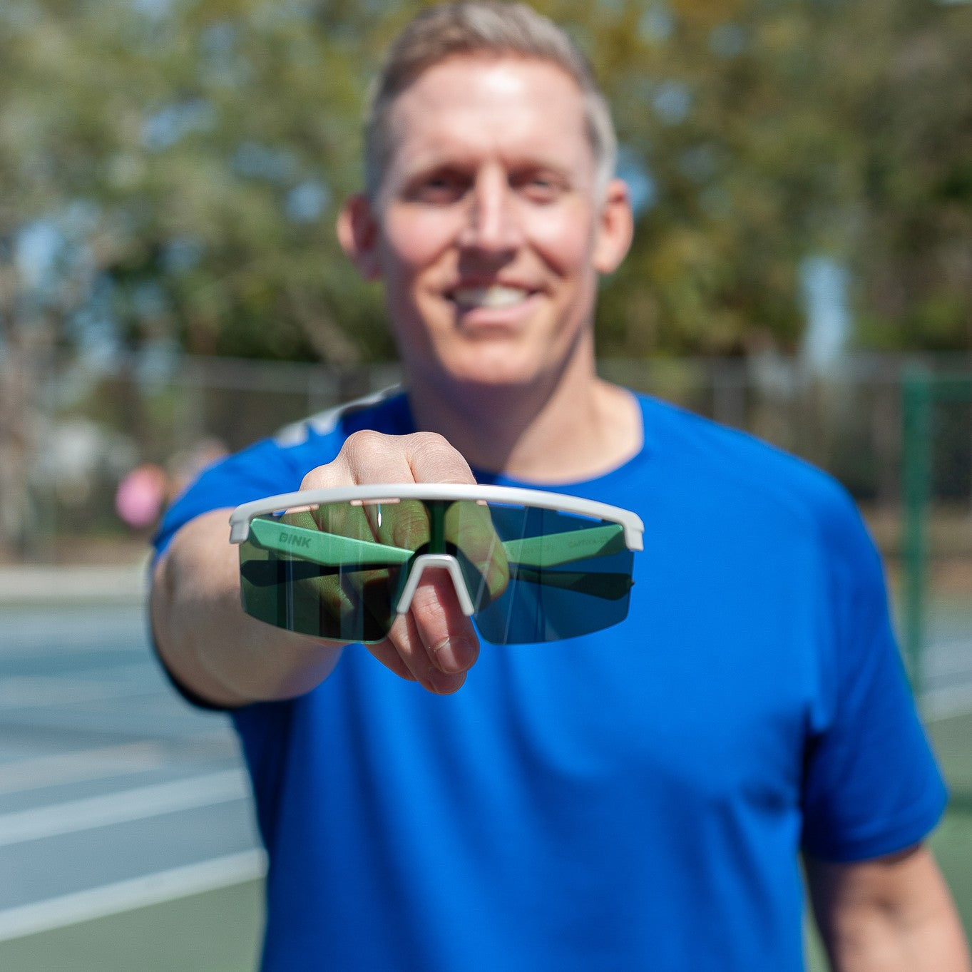 A man in a bright blue shirt stands on a tennis court, smiling and holding up Dink Eyewear's Captiva Large White Pearl Sunglasses with ColorBoost lens technology toward the camera, with blurred trees in the background.