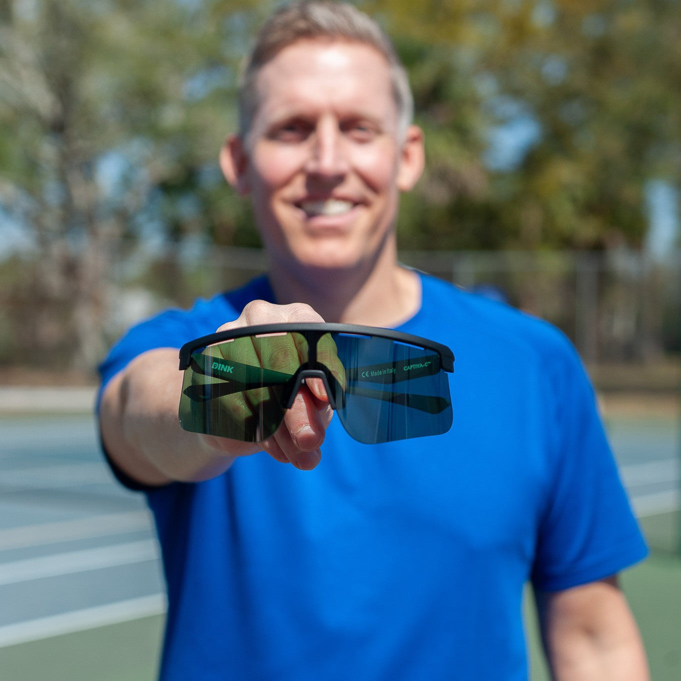 A man on an outdoor tennis court holds Dink Eyewear's Captiva Large Emerald Green Sunglasses with ColorBoost lens technology toward the camera, focusing on the eyewear. He wears a blue shirt, with trees and a net visible in the background.