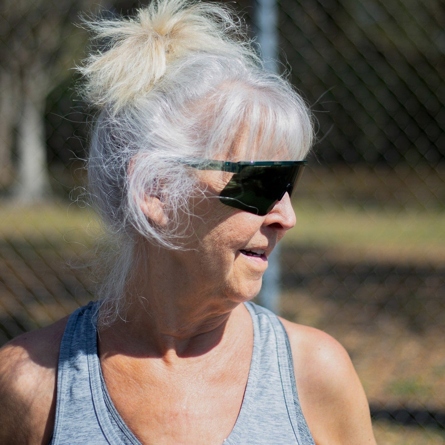 An older woman with light gray hair in a messy bun wears Dink Eyewear’s Captiva Emerald Green Pickleball Sunglasses with ColorBoost lenses and a gray tank top, smiling outdoors on a sunny day against a backdrop of greenery and a blurred fence.