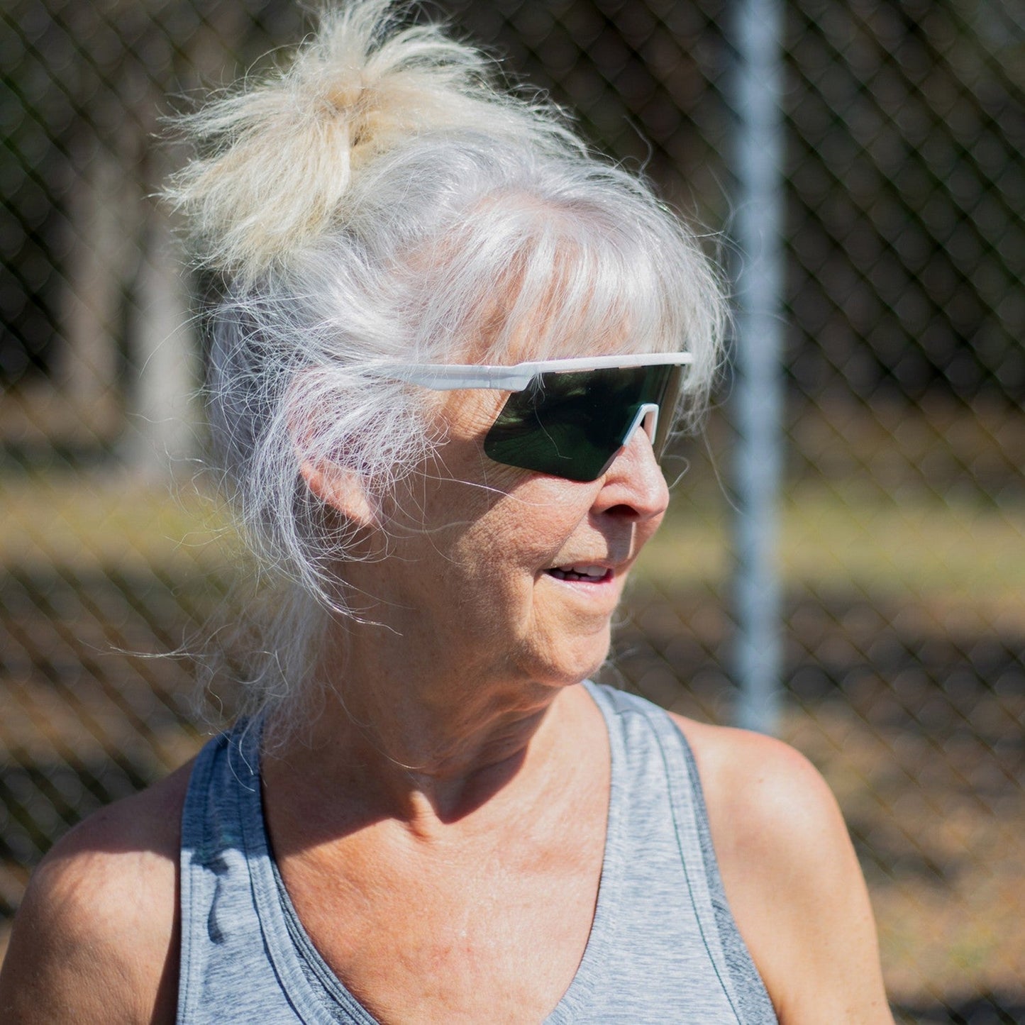 An older woman with long white hair in a ponytail smiles outdoors by a chain-link fence, wearing a light gray tank top and Dink Eyewear's Captiva Pickleball Sunglasses Performance Sport Eyewear with UV Protection. Sunlight highlights her face and hair.