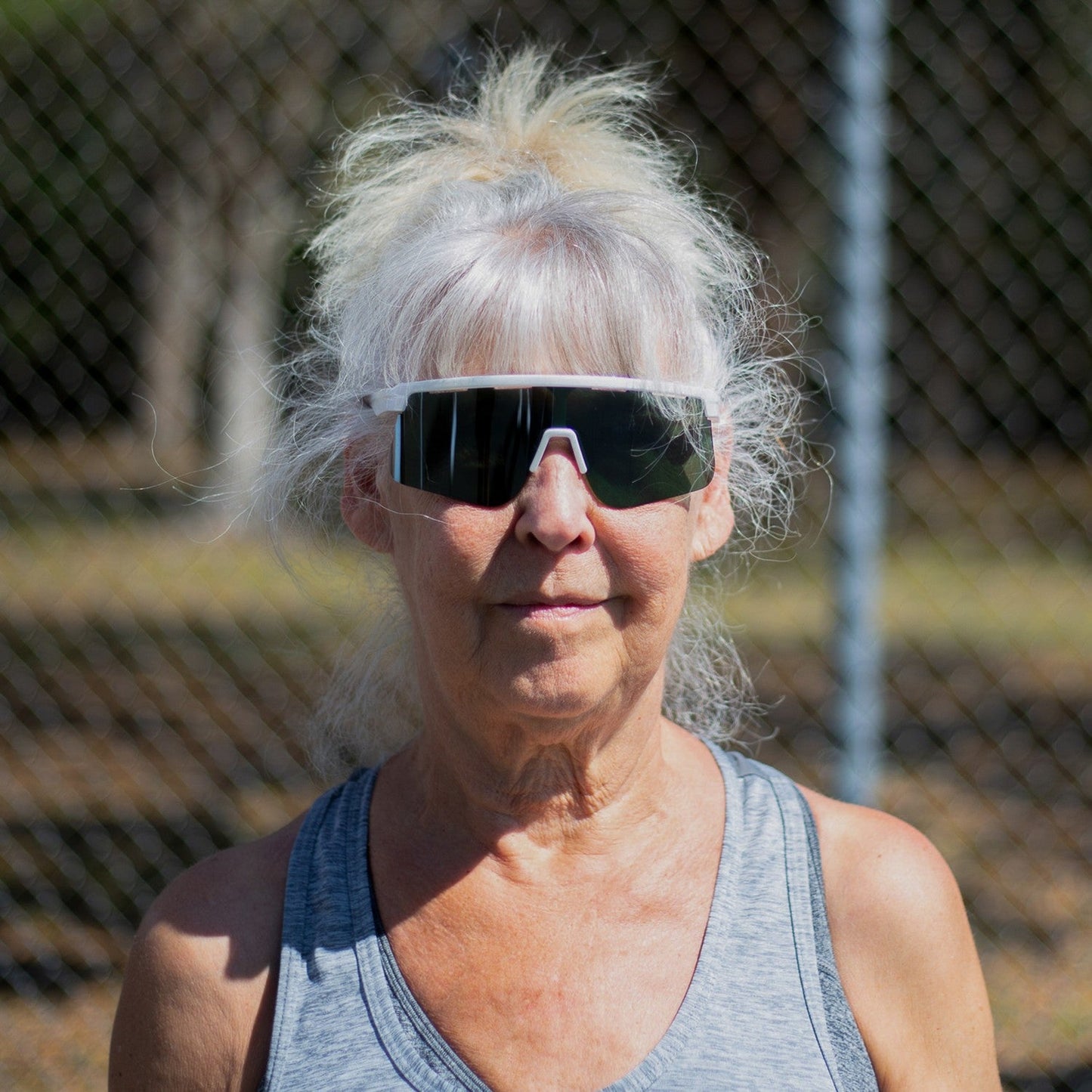 An older woman with long white hair in a ponytail wears Dink Eyewear's Captiva White Pearl Sunglasses and a gray tank top outdoors, with sunshine, greenery, and a blurred chain-link fence in the background.