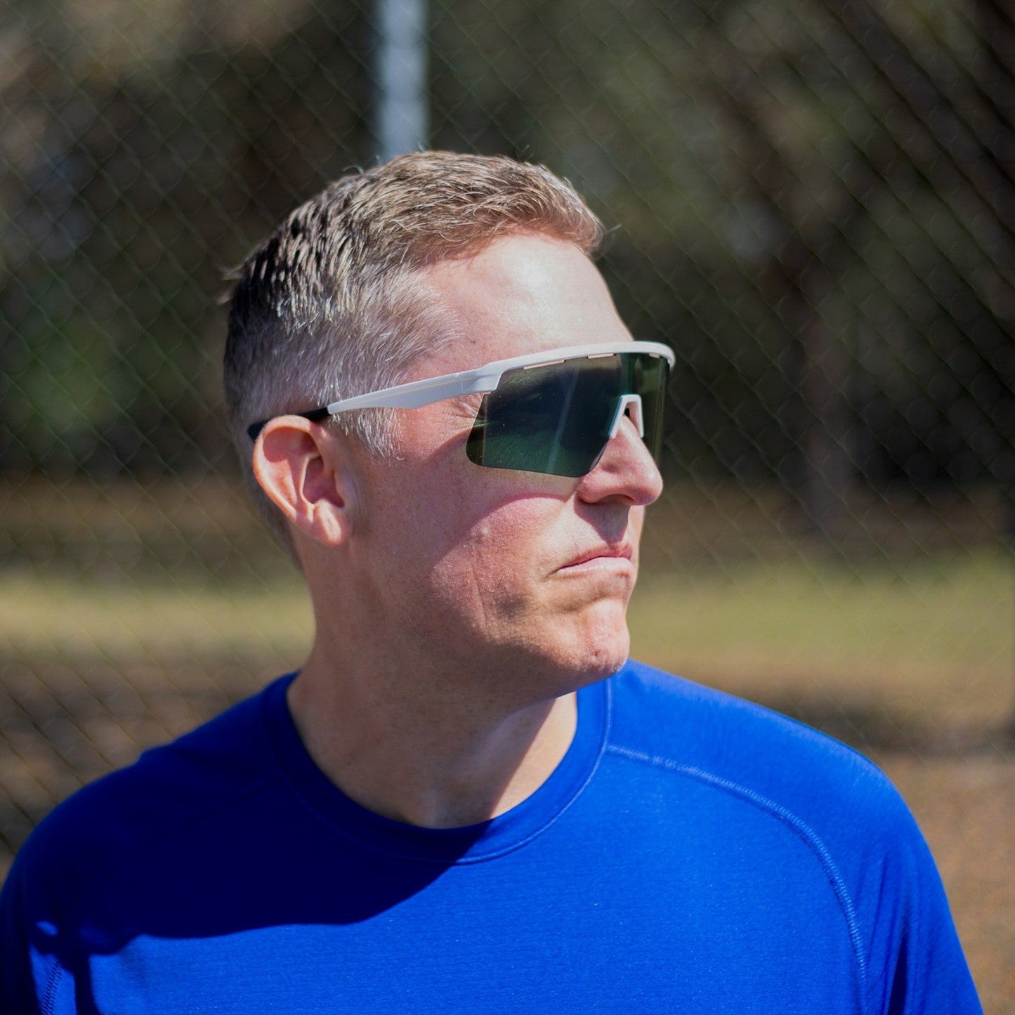 A man with short, light brown hair wears Dink Eyewear Captiva Pickleball Sunglasses with ColorBoost lens technology and UV protection. He sports a bright blue shirt outdoors on a sunny day, with trees and a fence blurred in the background.