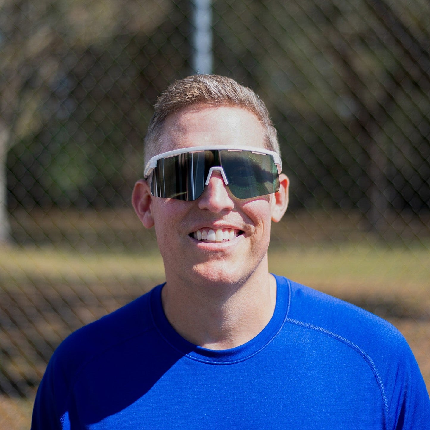 A smiling man with short light hair wears Dink Eyewear's Captiva Large White Pearl Sunglasses and a bright blue athletic shirt. He stands outdoors in front of a blurred chain-link fence, sunlight illuminating his face.