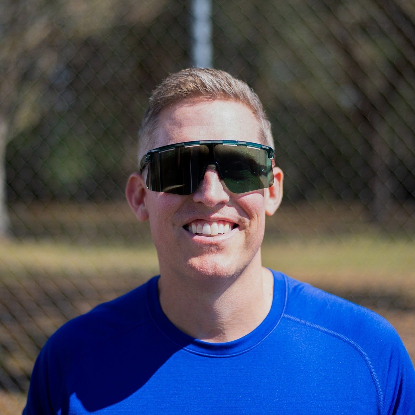 A man with short light brown hair, wearing Dink Eyewear’s Captiva Pickleball Sunglasses Performance Sport Eyewear with UV protection and a bright blue shirt, smiles outdoors near a blurred chain-link fence and trees in daylight.