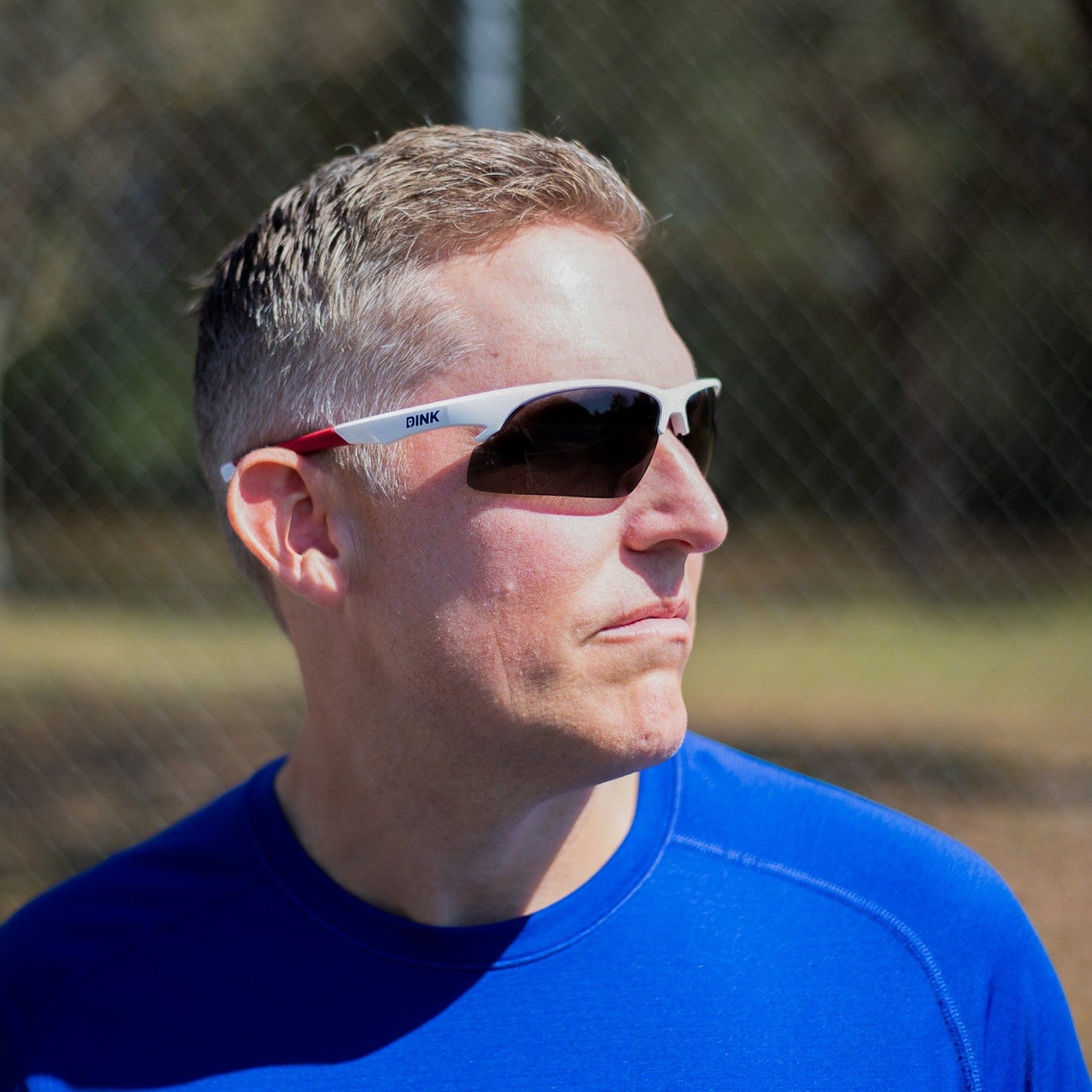 A man with short, light brown hair wears Dink Eyewear Daytona Sunglasses in White/Red and a blue athletic shirt. He is outdoors in sunlight, looking right, with a blurred chain-link fence and trees behind him.