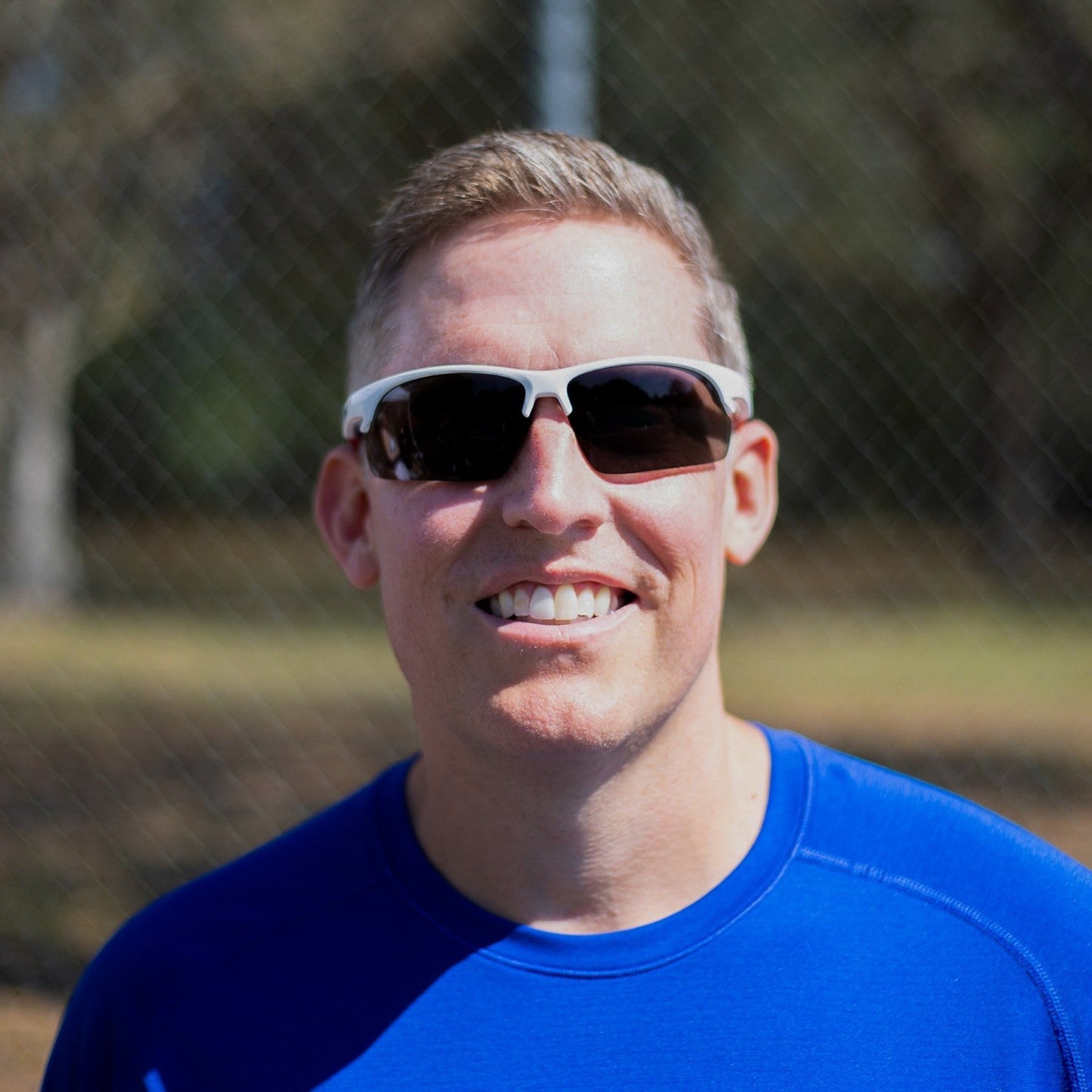 A smiling man in a bright blue shirt wears Dink Eyewear's Daytona Pickleball Sunglasses (White/Red) outdoors. The background is blurred, showing sunlight, trees, and a chain-link fence.