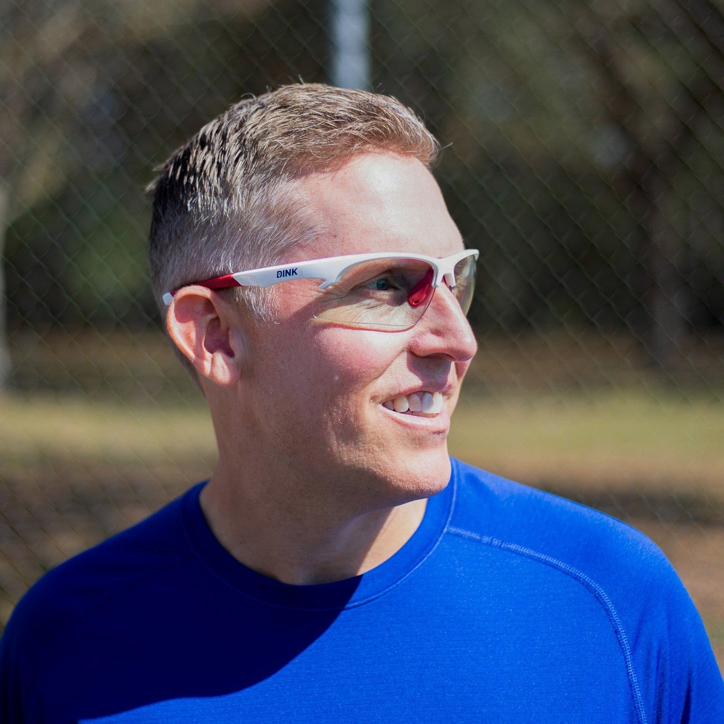 A smiling man with short light brown hair wears Dink Eyewear Daytona Sunglasses in White/Red and a royal blue athletic shirt. Outdoors by a chain-link fence, he looks to the side as sunlight highlights his face against a tree-filled background.