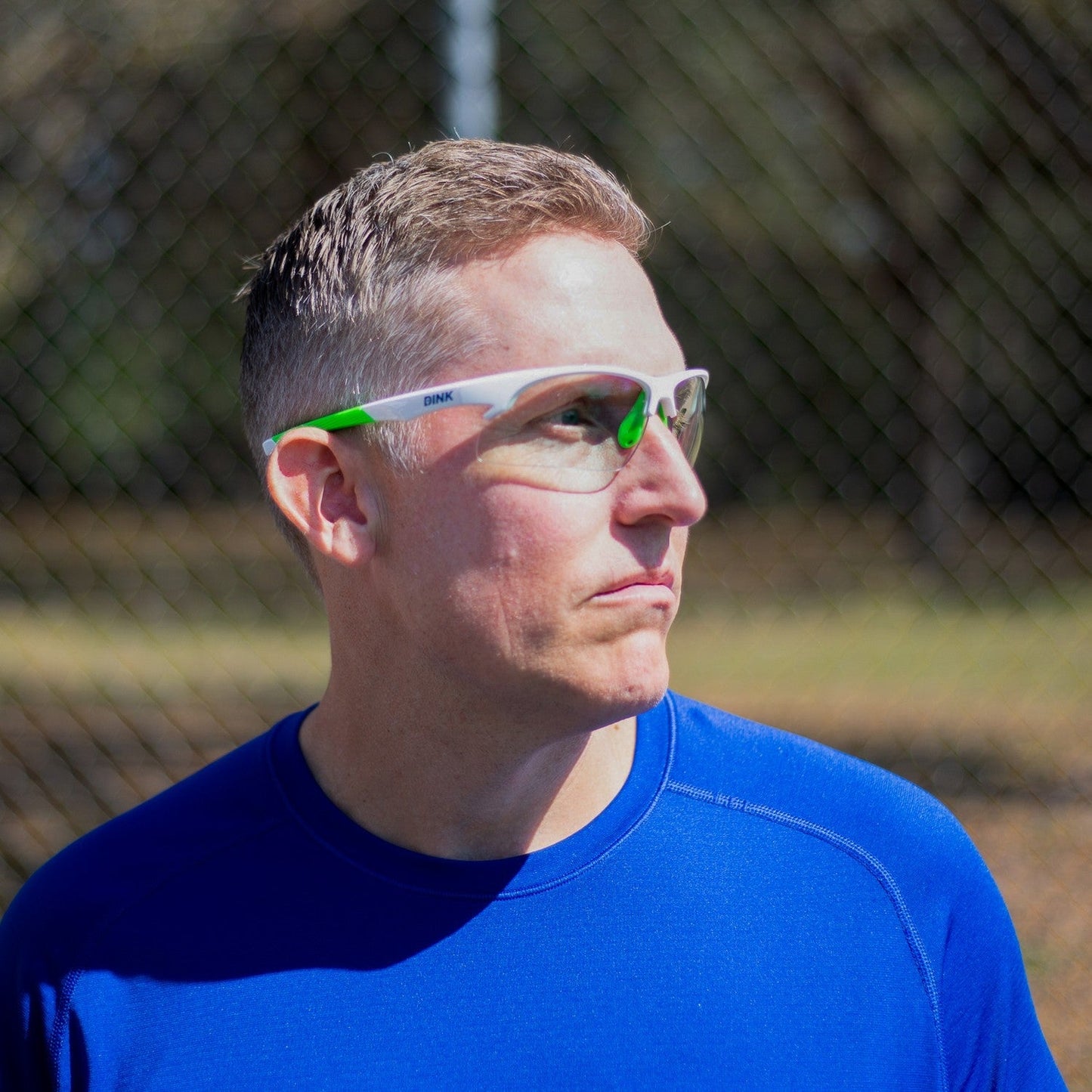 A man with short light brown hair wears Dink Eyewear's Daytona Pickleball Sunglasses - White/Green and a blue shirt outdoors by a chain-link fence, looking left with a neutral expression. Greenery appears blurred in the background.