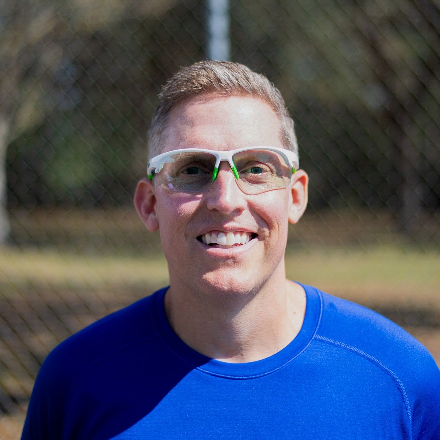 A smiling man in a bright blue shirt wears Dink Eyewear’s Daytona Sunglasses (White/Green) outdoors, with a sunlit park and blurred trees behind him—ideal for activities like pickleball.