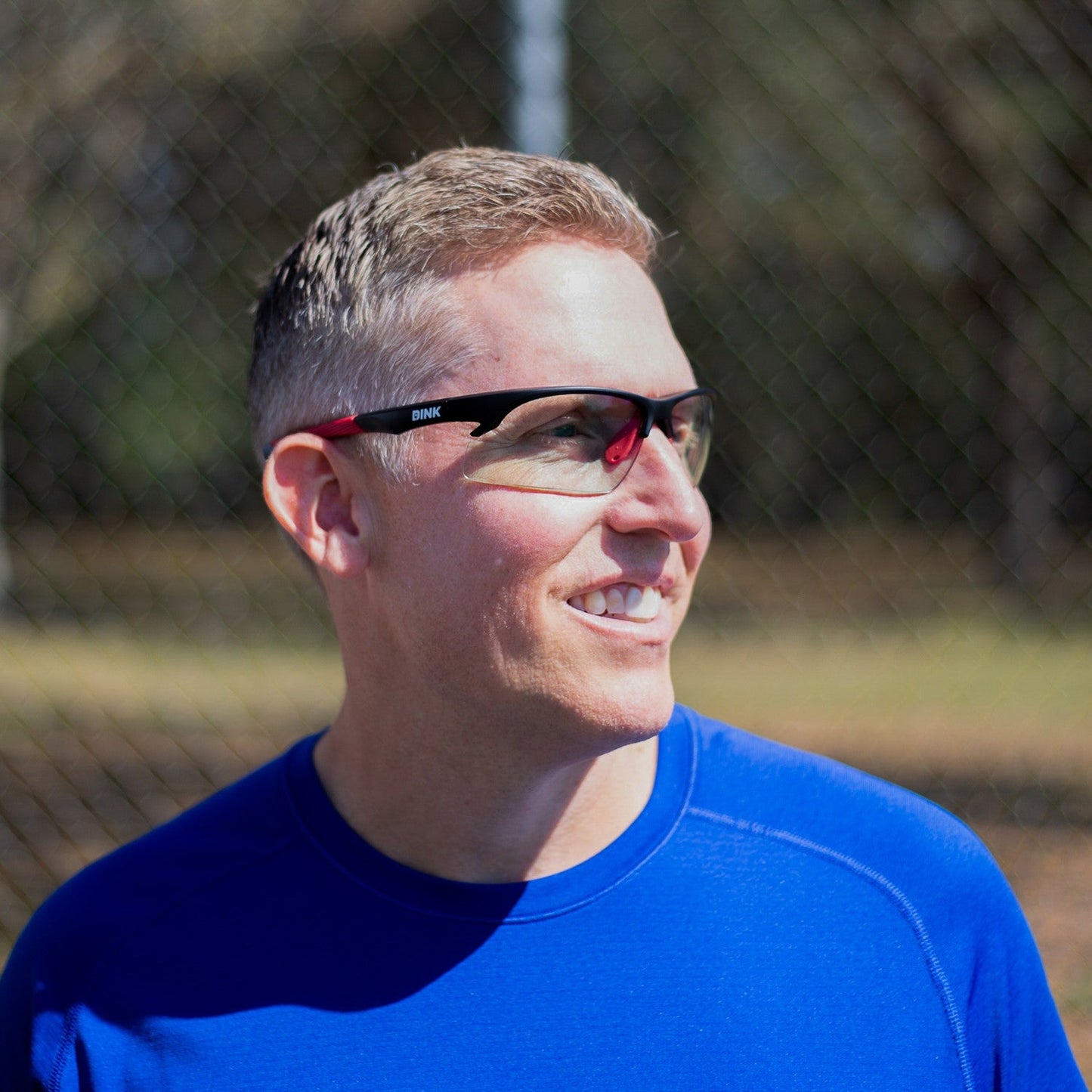 A man with short light brown hair, wearing Dink Eyewear Daytona Sunglasses in Black/Red and a bright blue shirt, stands outdoors smiling with a blurred chain-link fence and trees in the sunny background.