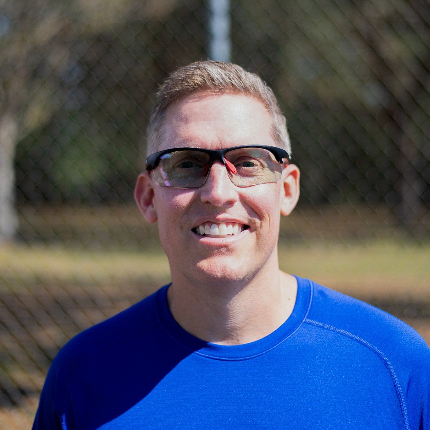 A man wearing Dink Eyewear's Daytona Sunglasses in Black/Red and a blue shirt stands outdoors, smiling at the camera. Sunlight, a blurred chain-link fence, and trees form the background. He has short light hair and black-rimmed frames.