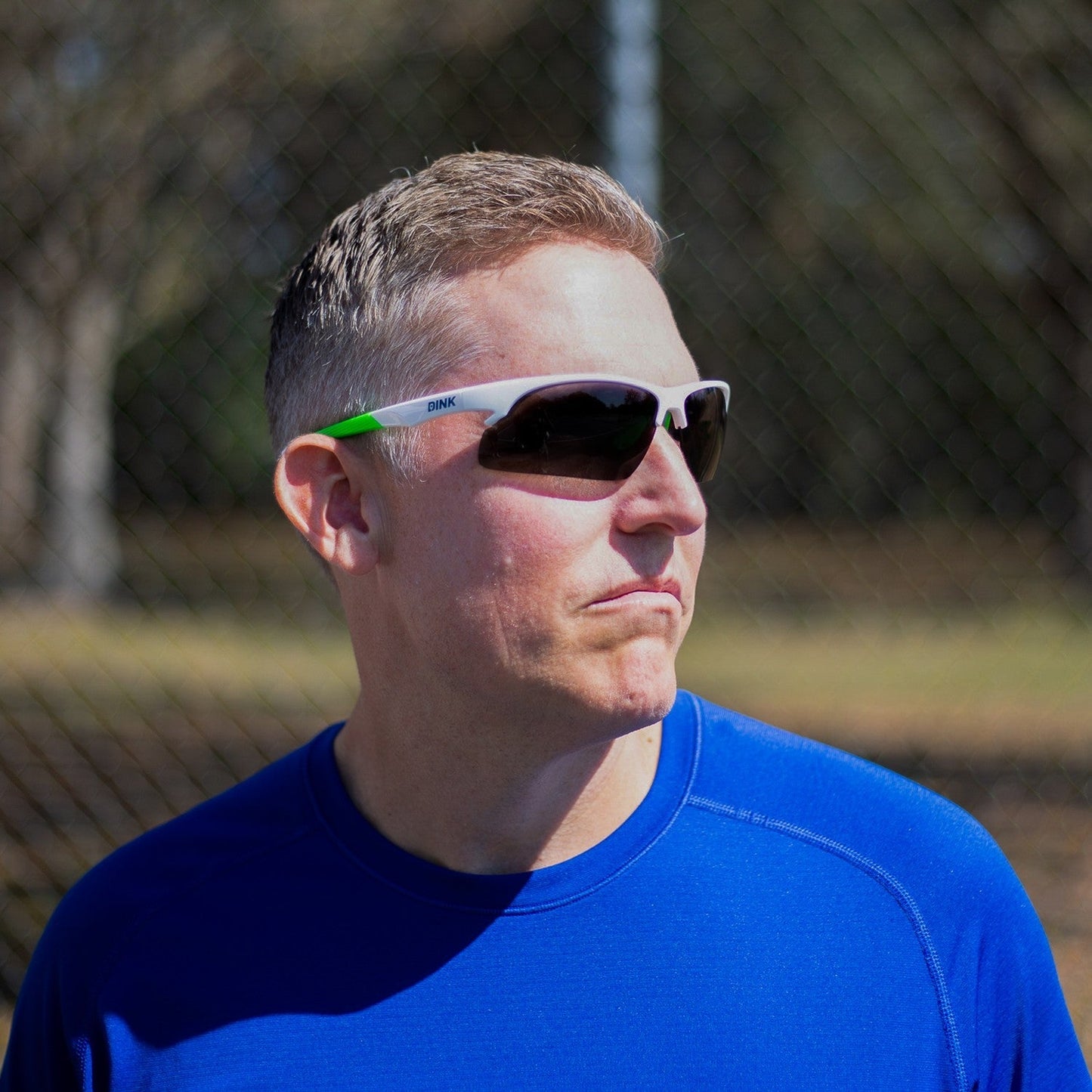 A man with short light brown hair stands outdoors in a royal blue athletic shirt, wearing Dink Eyewear's Daytona Pickleball Sunglasses in White/Green. A chain-link fence and trees suggest a sunny day at a pickleball park.