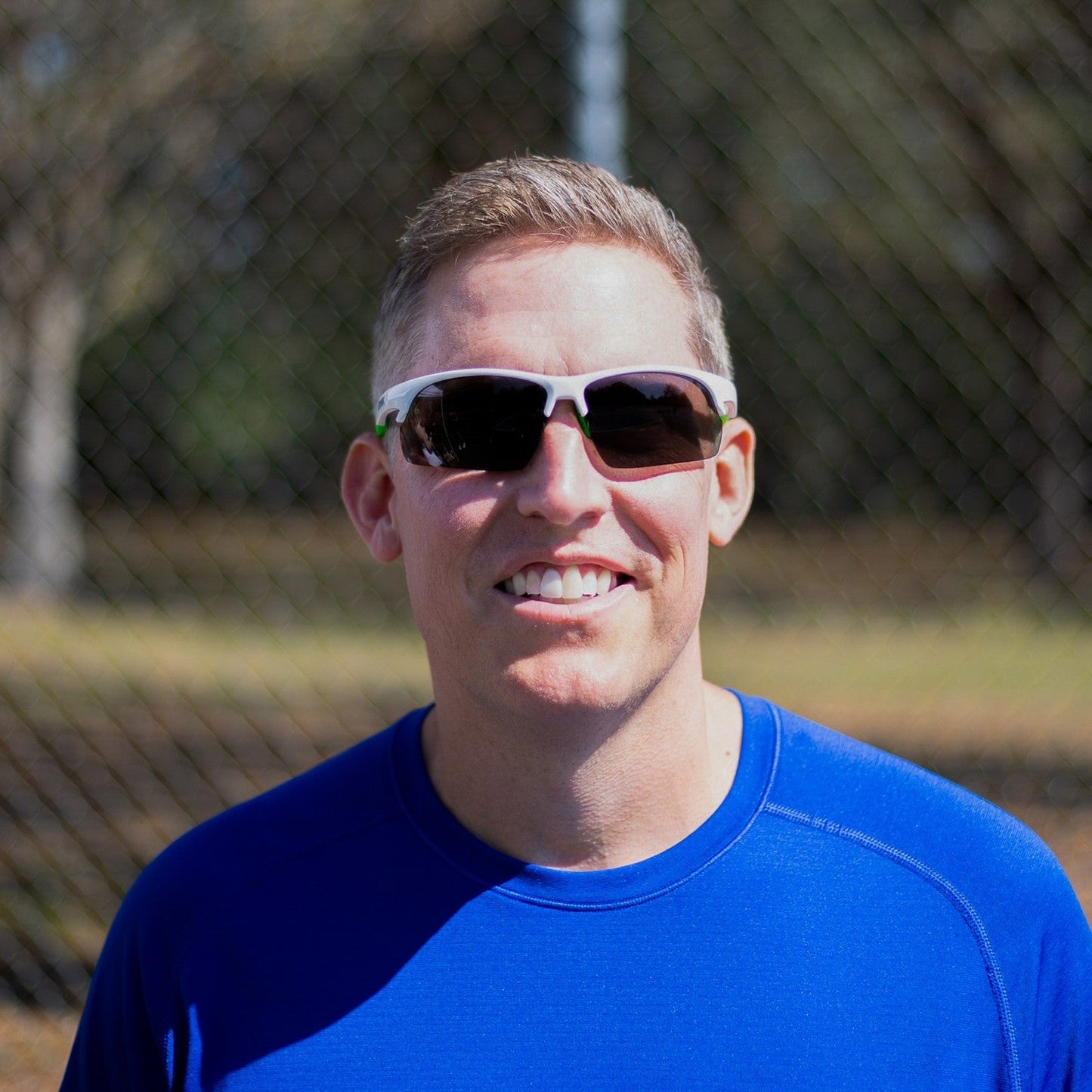 A man in a bright blue athletic shirt and short light brown hair smiles outdoors wearing Dink Eyewear Daytona Pickleball Sunglasses in White/Green. Sunlit trees and a blurred fence hint at an outdoor pickleball court.