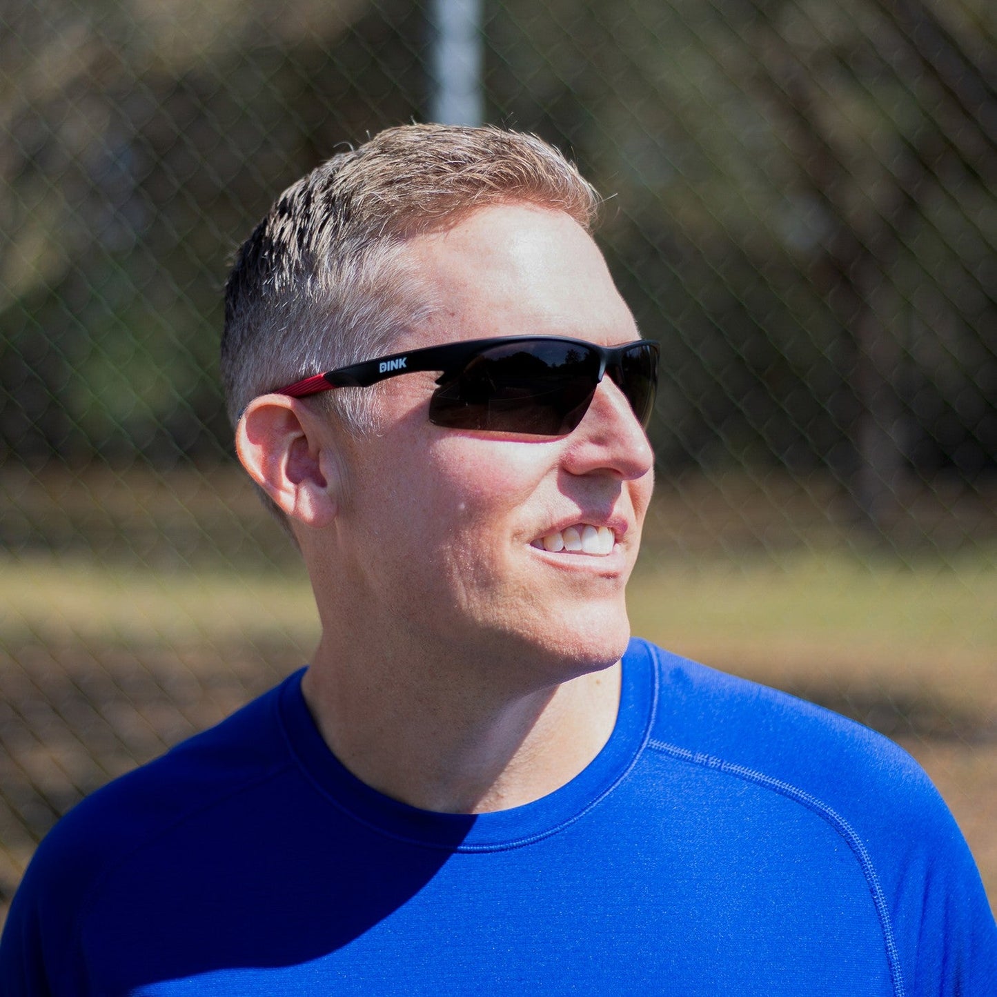 A man in a bright blue athletic shirt smiles while wearing Dink Eyewear's Daytona Sunglasses in Black/Red. He stands outdoors with short light brown hair, against a backdrop of trees and a blurry chain-link fence on a sunny day.
