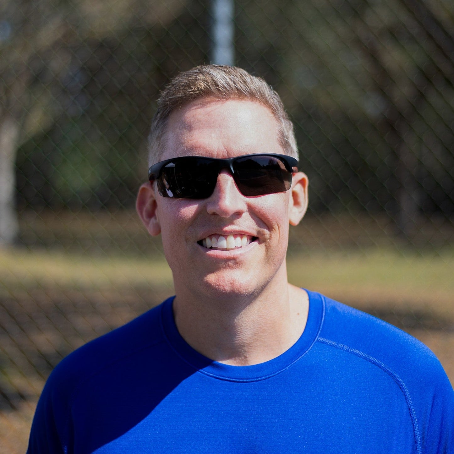 A smiling man with short light hair wears Dink Eyewear Daytona Sunglasses in Black/Red and a bright blue shirt while standing outdoors in sunlight, with a blurry chain-link fence and trees in the background.