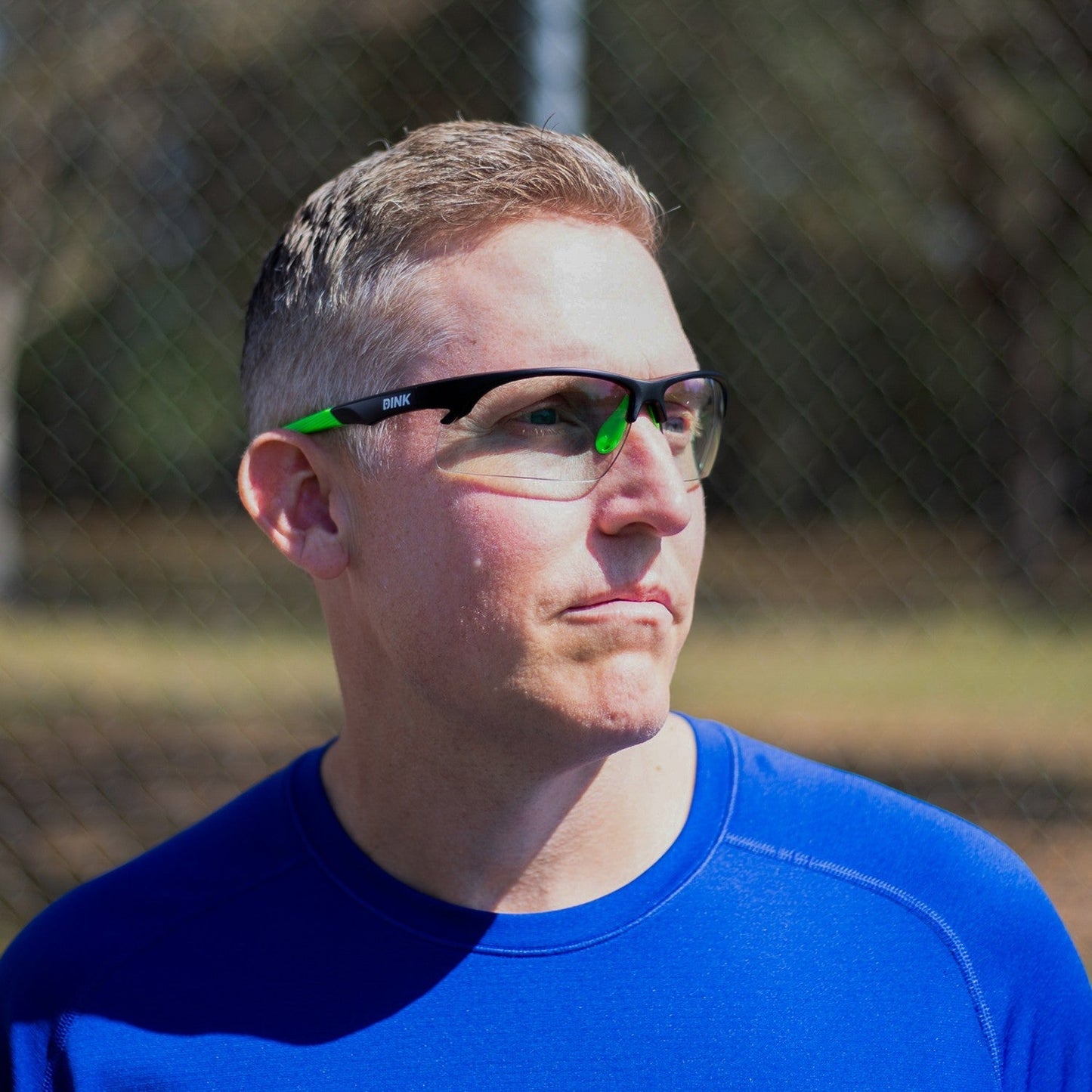 A man with short blond hair wears Dink Eyewear's Daytona Pickleball Sunglasses - Black Green and a blue shirt outdoors, looking to the side. Green trees and a blurred chain-link fence appear in the background.