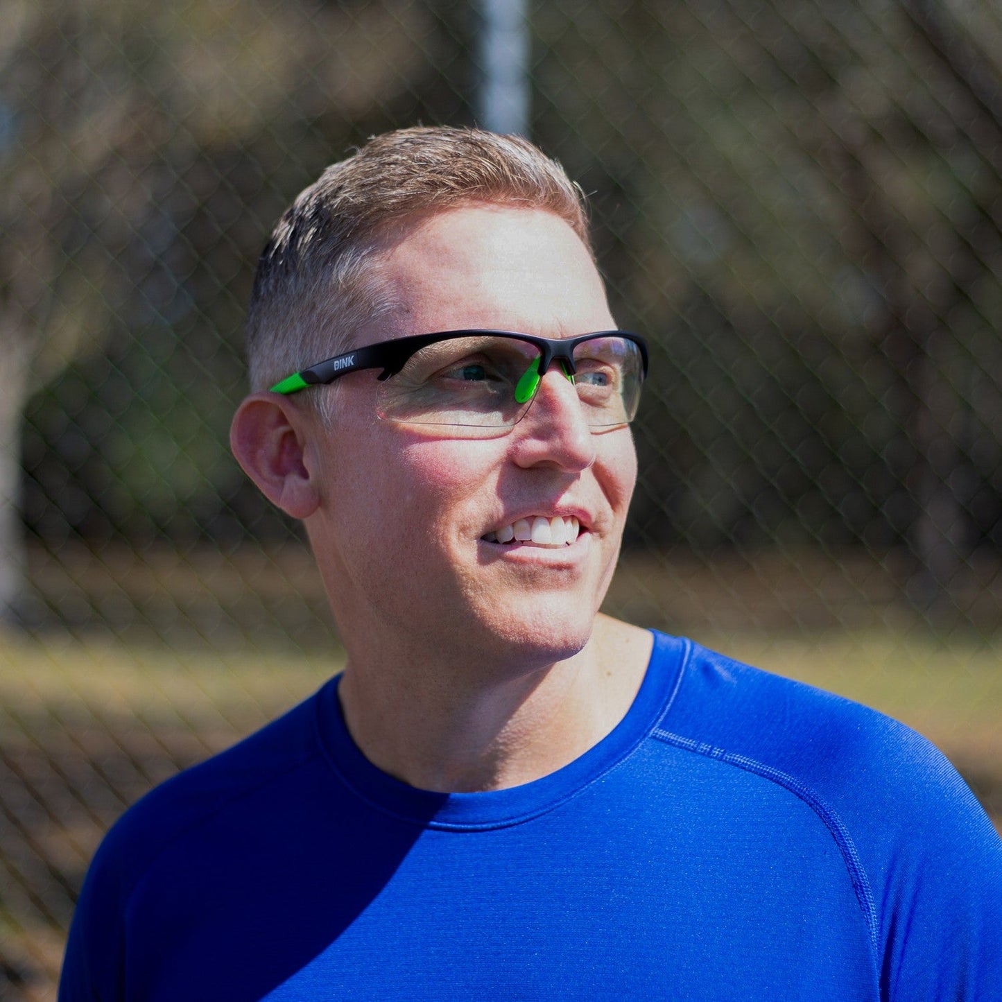 A man with short, light brown hair and light skin wears Dink Eyewear's Daytona Sunglasses in Black Green with a lightweight frame and a royal blue shirt, smiling as he looks left. A blurred wire fence and trees appear in the background.