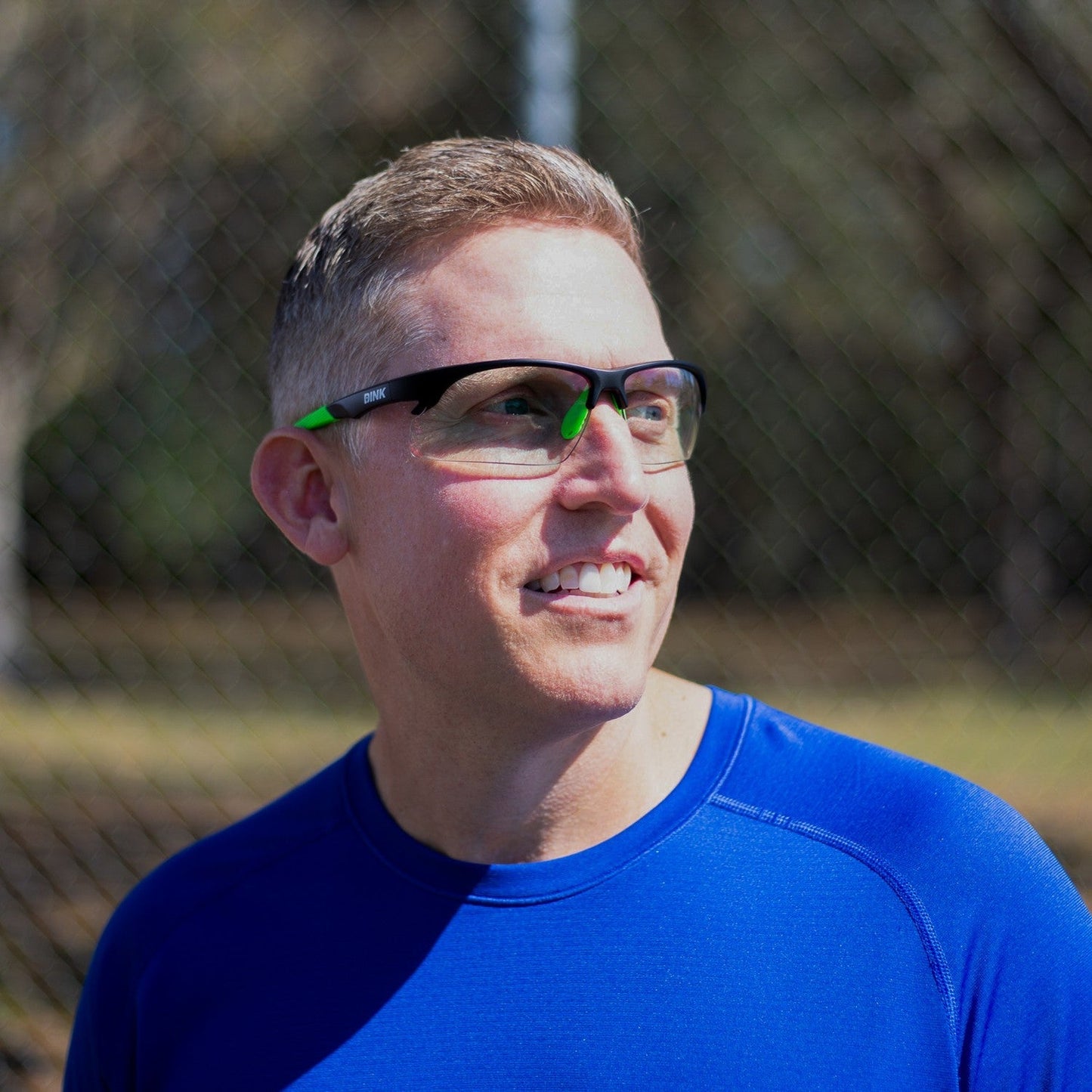A man in a bright blue athletic shirt smiles and looks to the side, wearing Dink Eyewear Daytona Pickleball Sunglasses - Black Green. He stands outdoors with sunlight, trees, and a blurry chain-link fence in the background.