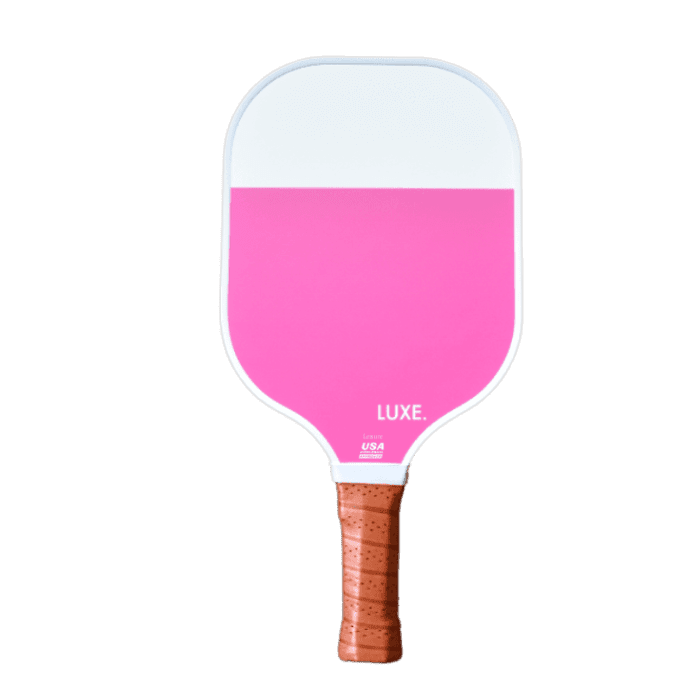 The LUXE Pickleball Dipped - Leisure paddle has a rounded rectangular shape with a bright pink fiberglass surface on the bottom two-thirds, white on the top third, and “LUXE.” printed near the bottom. The brown handle features textured grip tape.