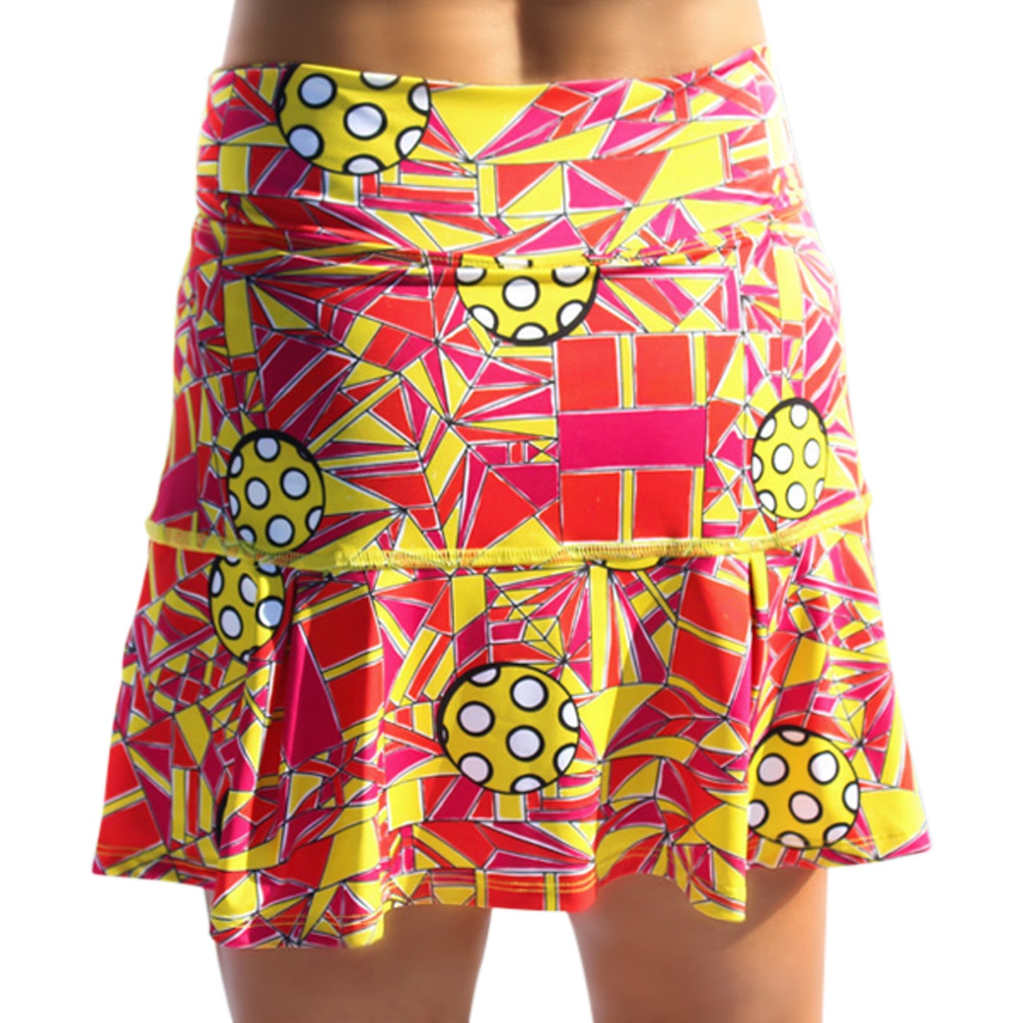 A person wears the Pickleball Bella PB Drop Pleat Skort Angle Shot 1, featuring bold red, yellow, and white geometric patterns. Its fitted waist, flared ruffled hem, and built-in compression shorts offer style and comfort.