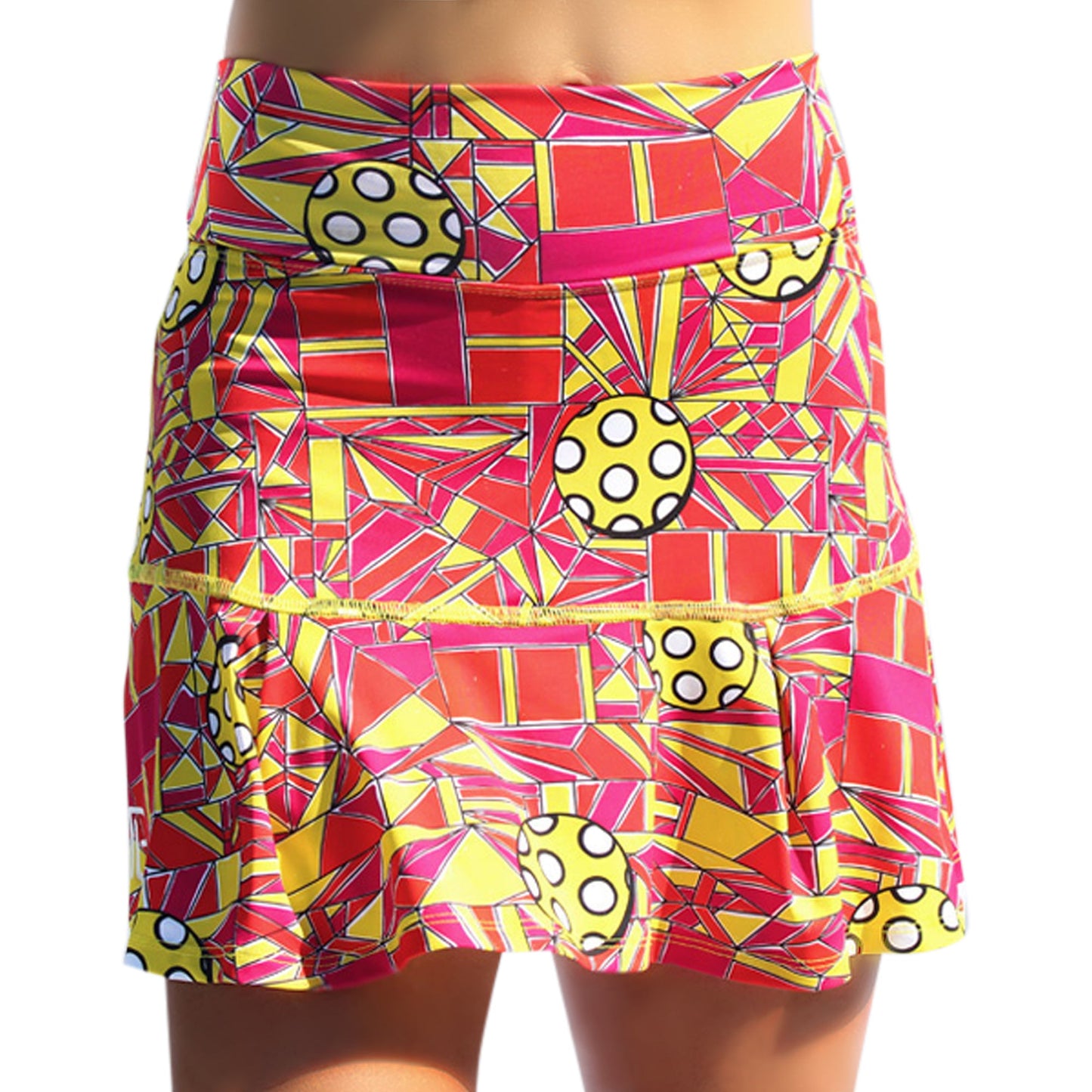 A person wears the Pickleball Bella PB Drop Pleat Skort Angle Shot 1, a brightly colored pickleball skirt with red, yellow, and orange geometric patterns, white circles with black dots, various shapes, and a slight hem ruffle.
