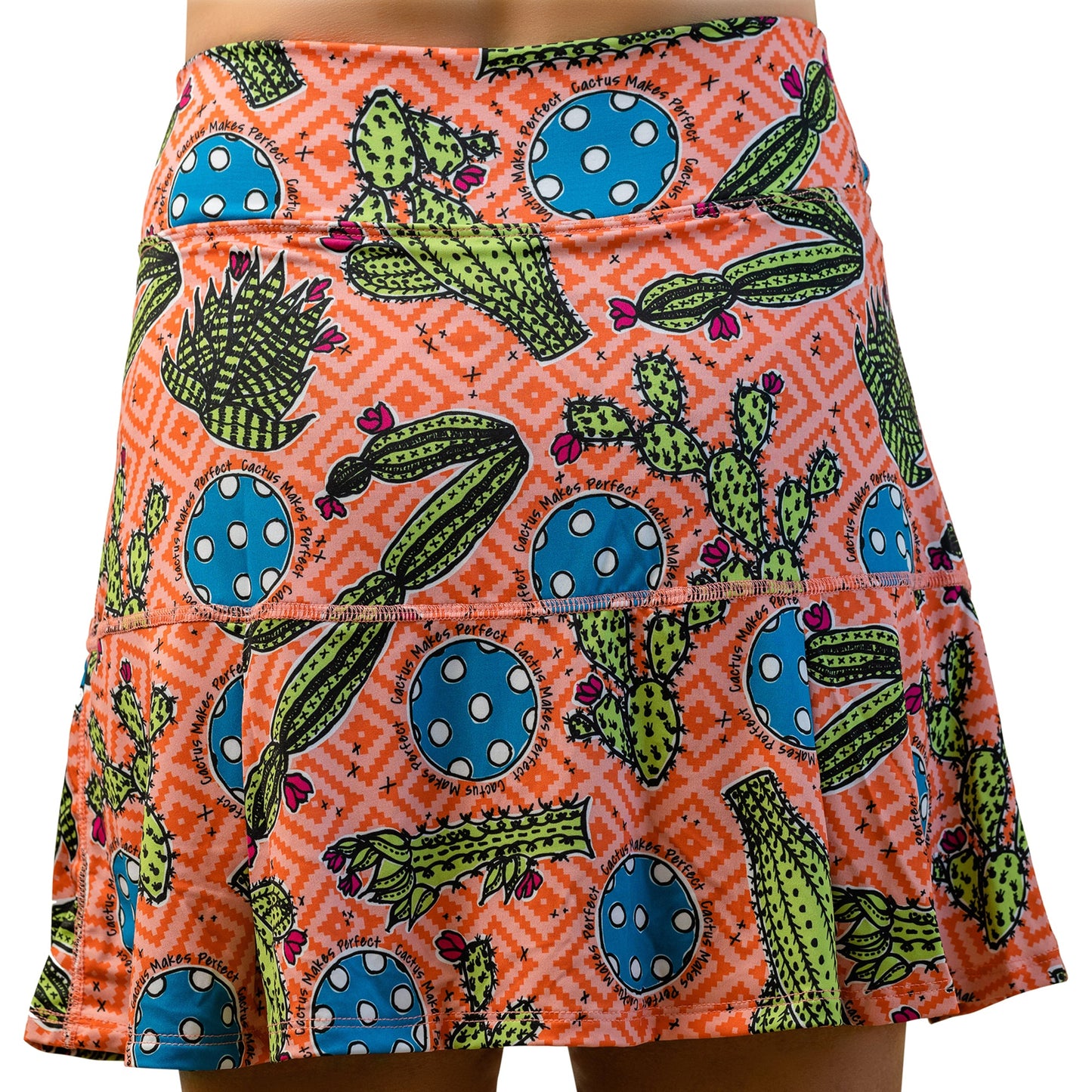 A person wearing the Pickleball Bella PB Drop Pleat Skort Cactus Makes Perfect 1—a short, orange athletic skirt with a southwestern print featuring green cactus, blue circles with white dots, pink geometric patterns, and a flared above-the-knee hem.