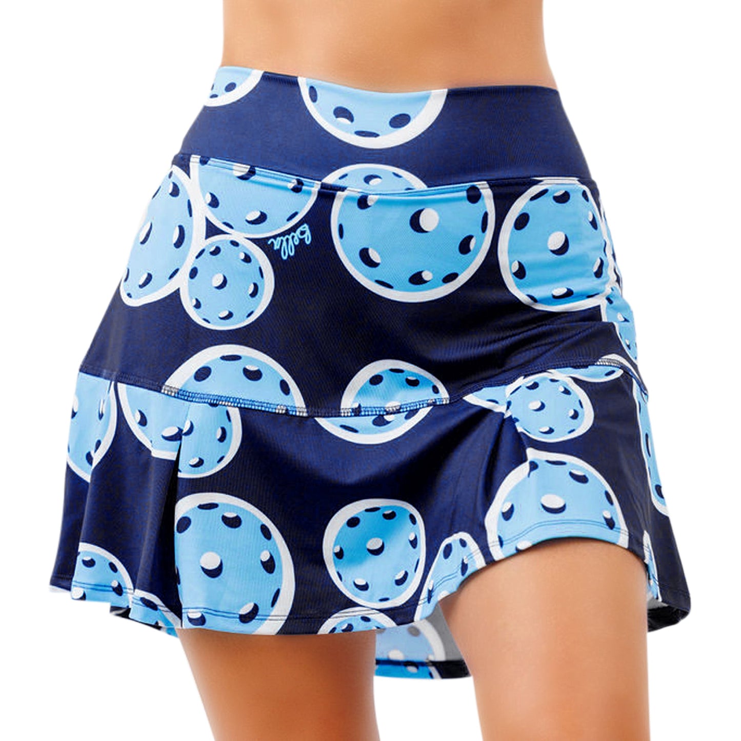 A woman wears the Pickleball Bella PB Drop Pleat Skort Double Bounce, a navy athletic skort with large light blue pickleball prints. This 4-way stretch activewear features a no roll waistband, tiered hem, and a flattering, playful fit.