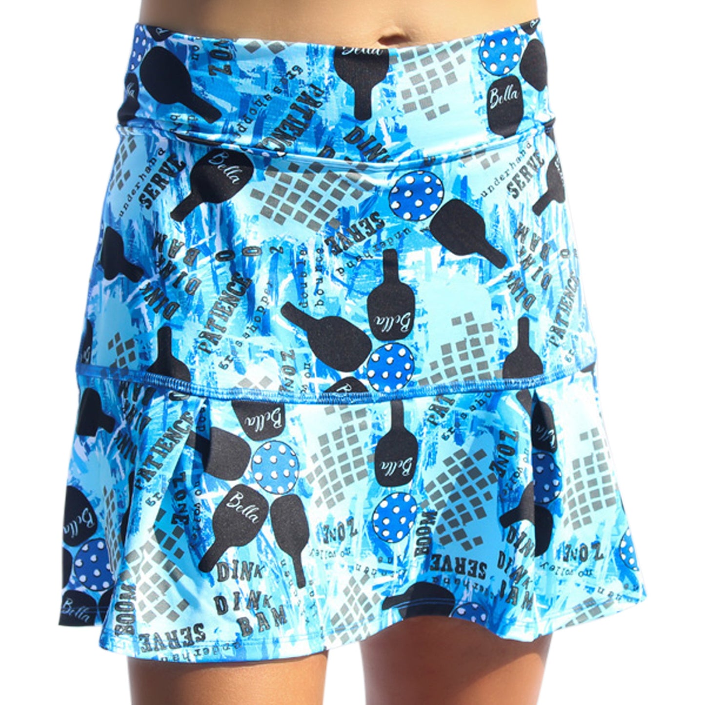 Pickleball Bella’s PB Drop Pleat Skort Graffiti 2 is a women’s athletic skirt with a blue and white graffiti print, black and blue pickleball paddles, polka-dot balls, text like “DINK” and “SERVE,” a fitted waistband, and ruffled hem.