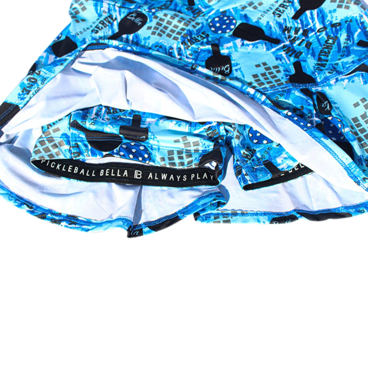 The Pickleball Bella PB Drop Pleat Skort Graffiti 2 features blue graphic prints with paddles, balls, and geometric shapes, plus a built-in white liner and an inner waistband that reads "PICKLEBALL BELLA ALWAYS PLAY.