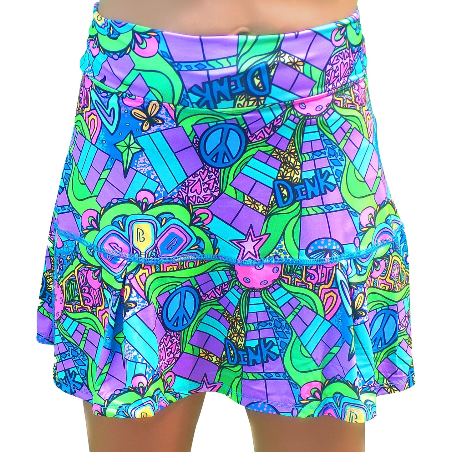 The PB Drop Pleat Skort Groovy by Pickleball Bella features a vibrant psychedelic retro print in blue, green, pink, and purple with peace signs, stars, hearts, and "Denk" on its ruffled layers. Modeled by someone with tan skin.
