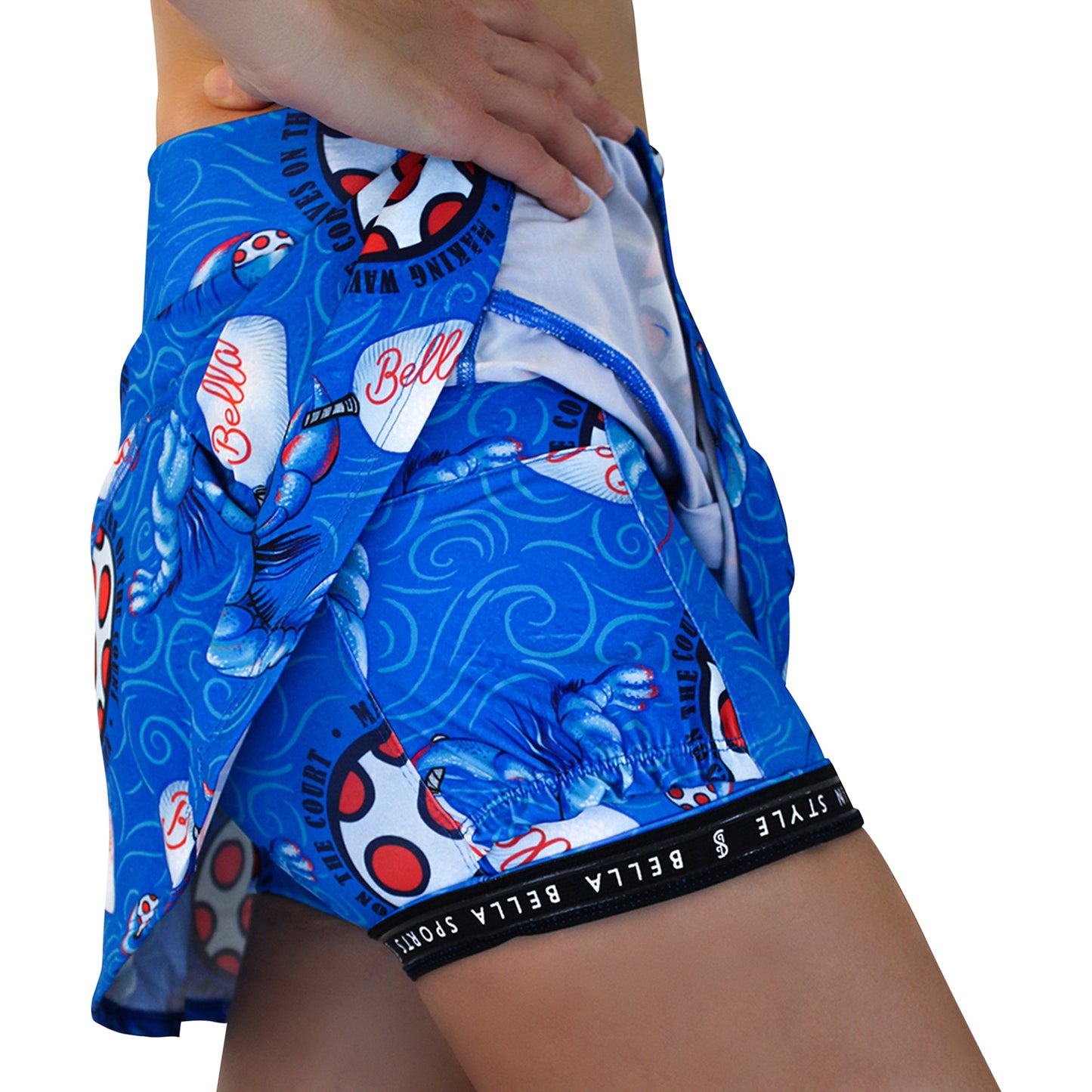 A close-up of a person wearing the Pickleball Bella PB Drop Pleat Skort Lobster Monster, featuring a blue, red, and white graphic design with built-in white underwear trimmed in blue. Only the lower torso and upper thighs are visible.