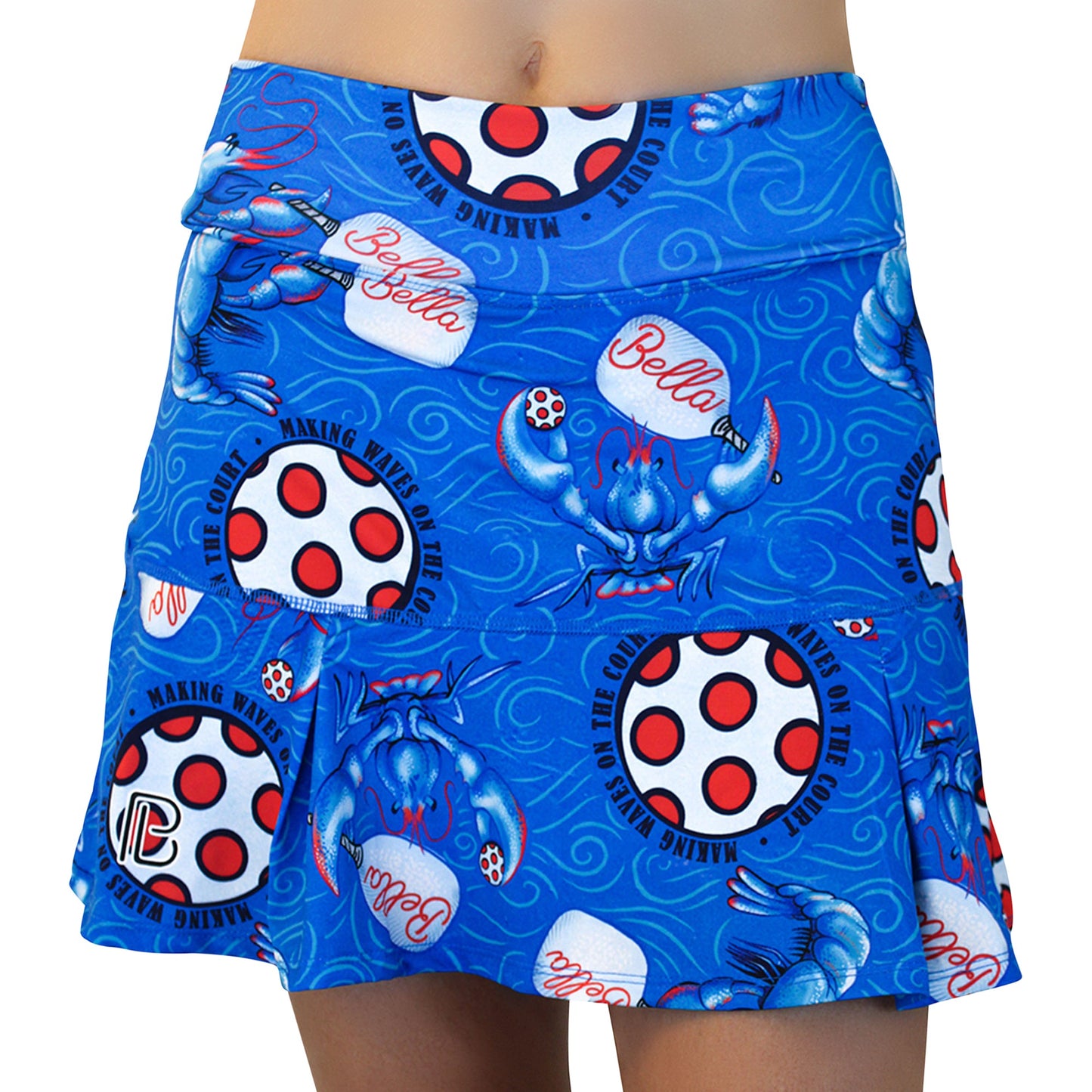The Pickleball Bella PB Drop Pleat Skort Lobster Monster features playful blue crabs, polka-dotted circles, “Bella,” and “Making Waves On The Course” text with wave swirls in a sporty, blue pleated design. Part of wearer’s midsection and legs visible.